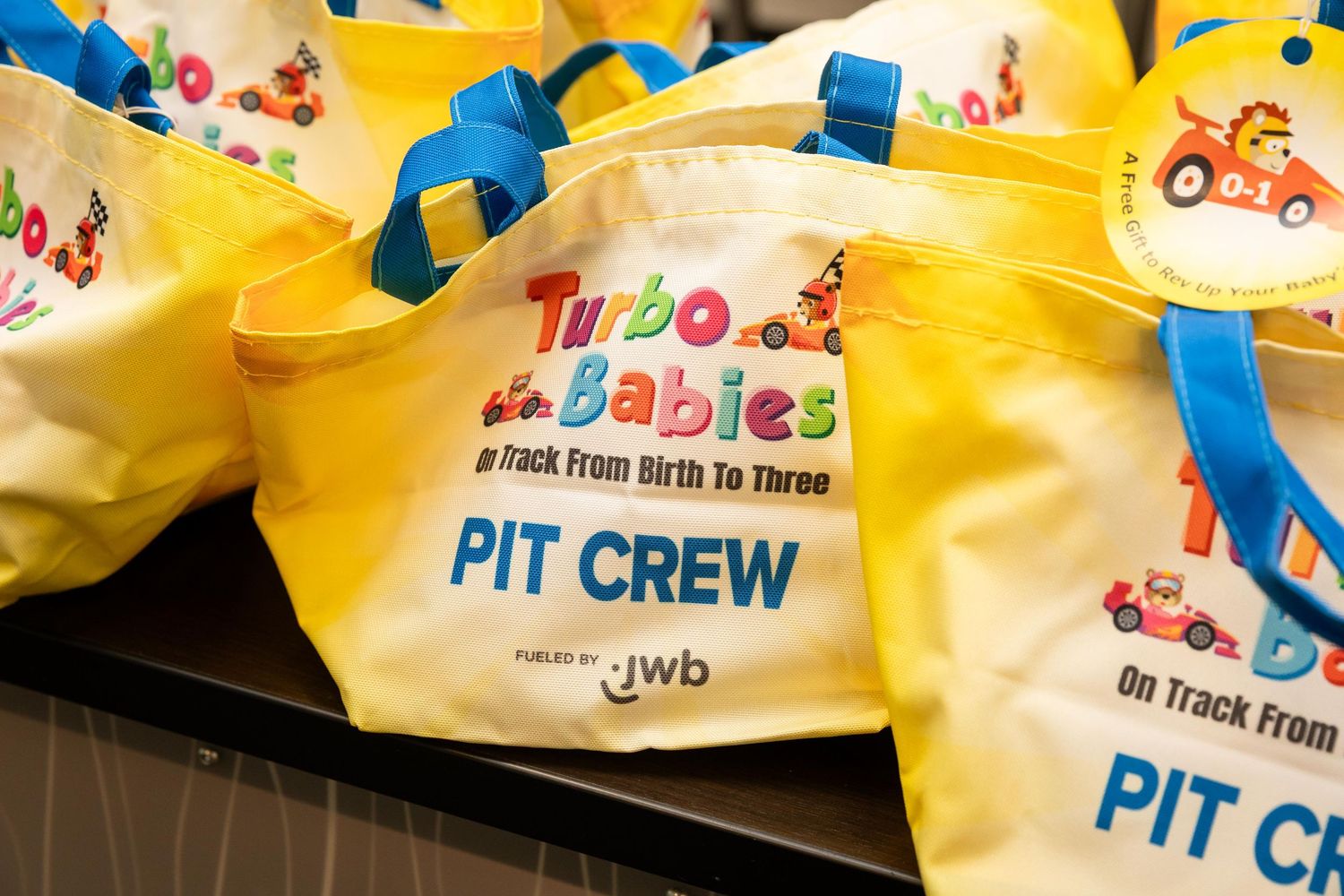 Yellow Turbo Babies Pit Crew tote bags with blue handles promoting a birth to three program by JWB.