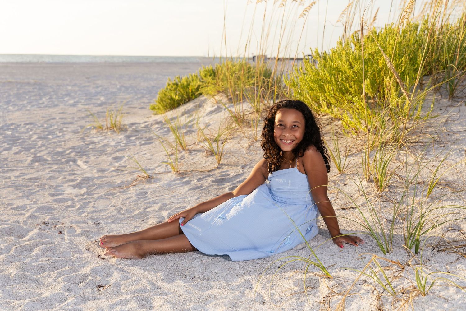 A smiling young girl in a light blue dress sits on sandy beach near sea oats and dunes on a sunny day.