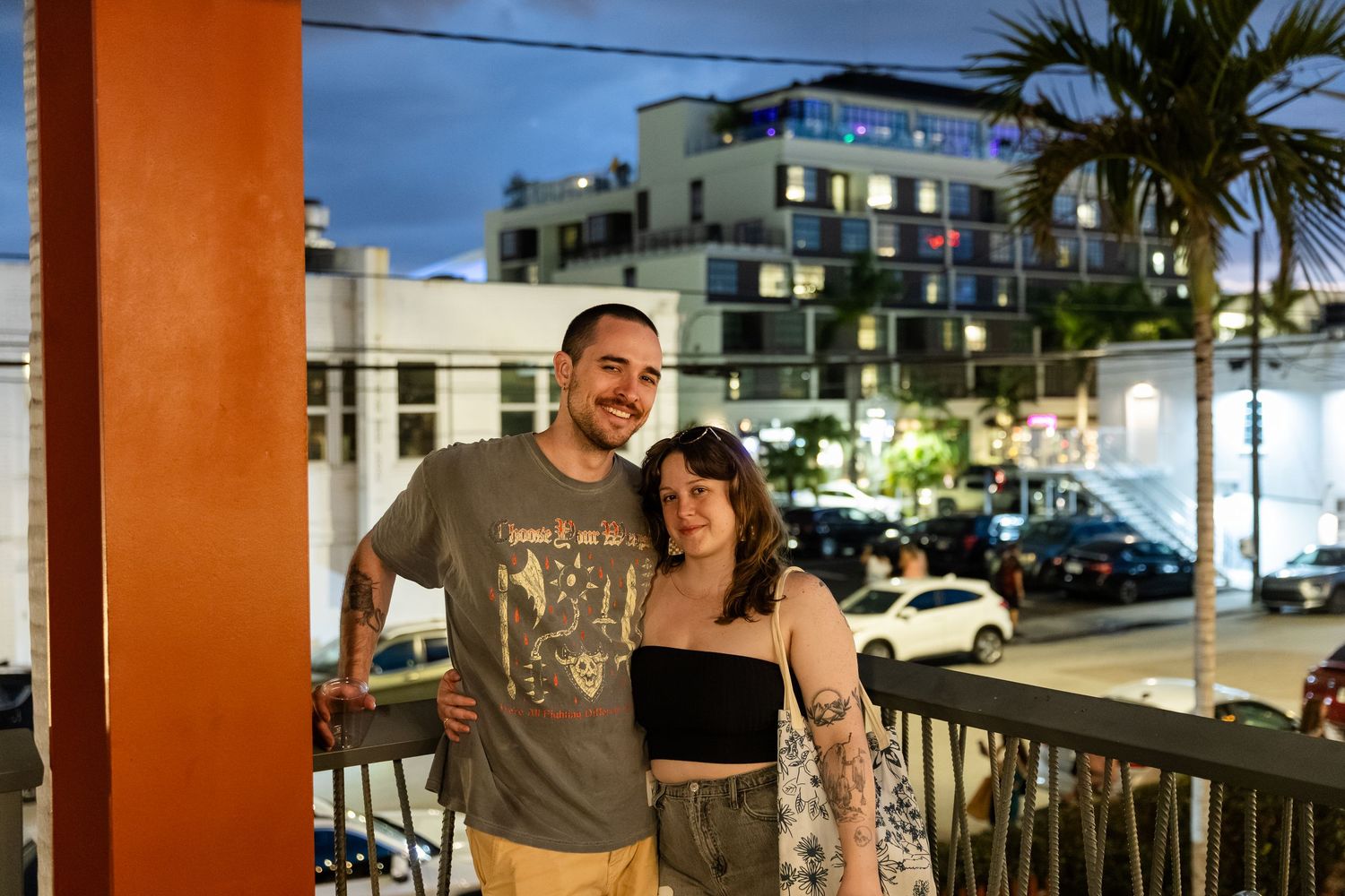A smiling couple with tattoos posing on a balcony at night with city lights behind them.