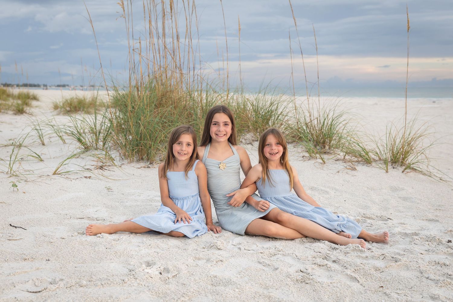 Three girls in matching blue dresses sitting together on white sand beach with sea oats and cloudy sky.