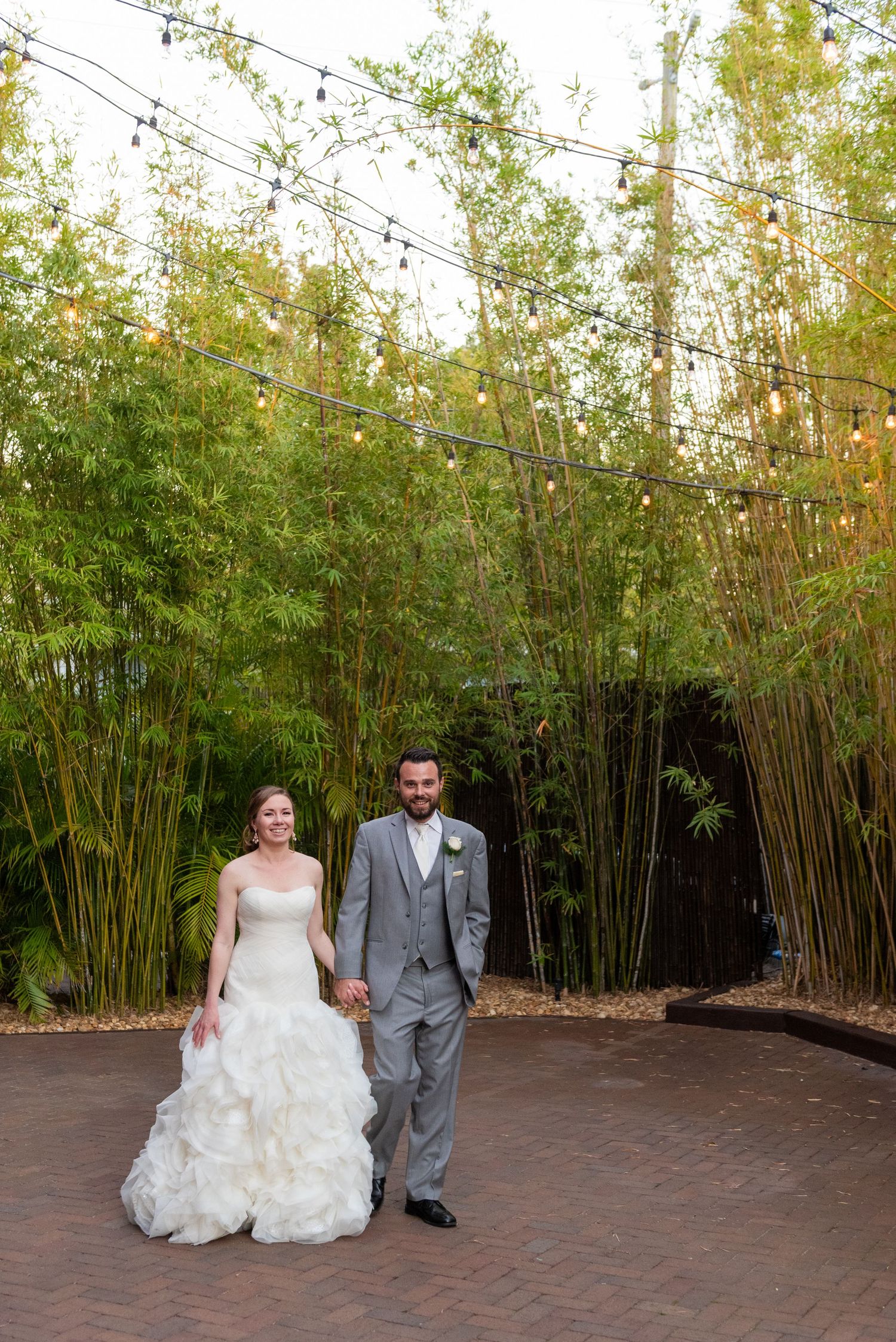 Smiling bride and groom walk hand-in-hand through bamboo garden with string lights overhead.