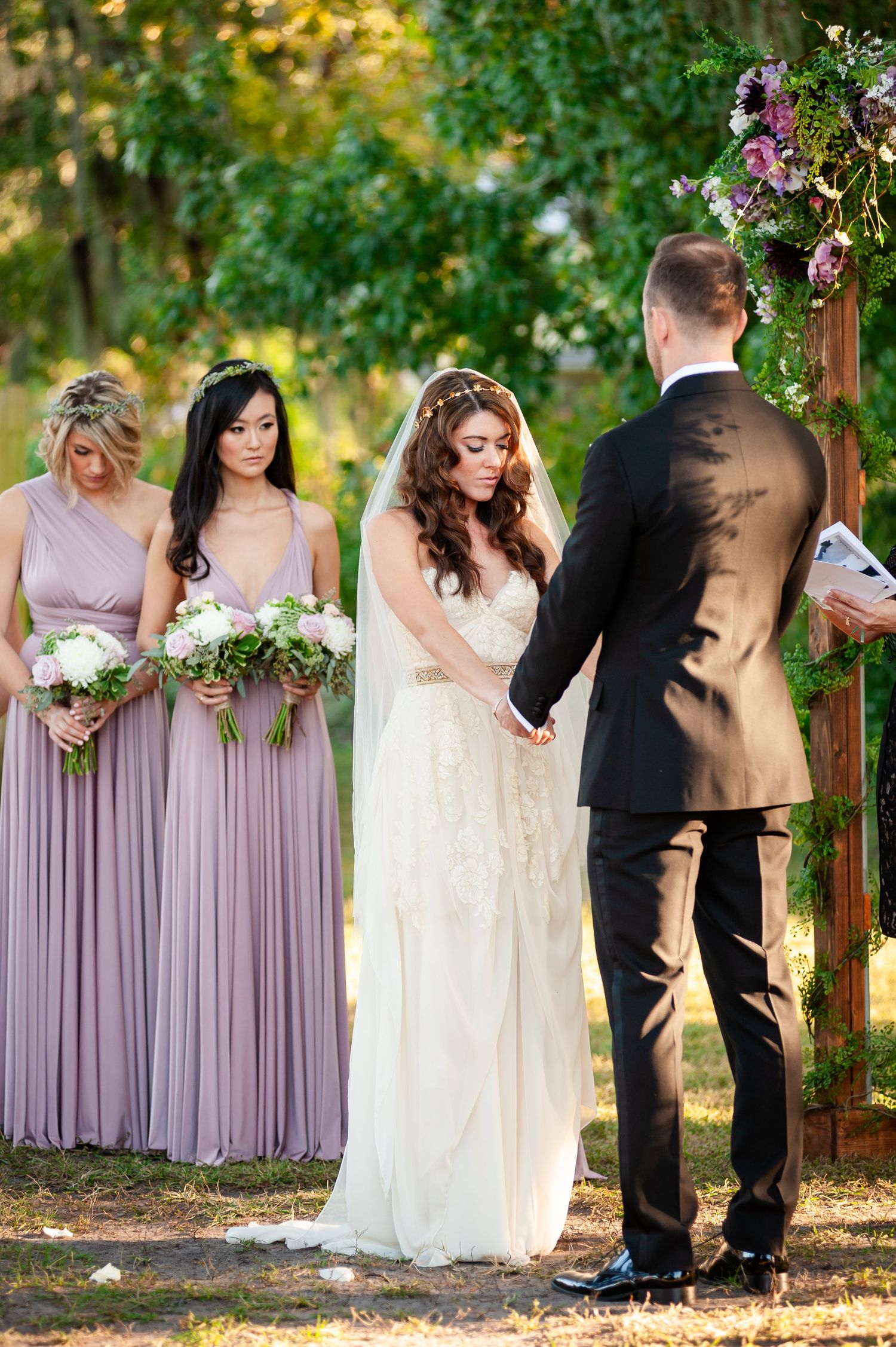 Bride and groom exchange vows as bridesmaids in lavender gowns hold white bouquets at outdoor ceremony.