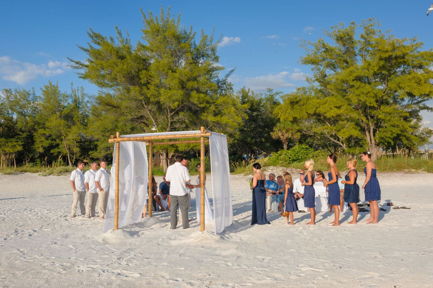 Wedding party stands at bamboo arch on sandy beach surrounded by lush green trees.