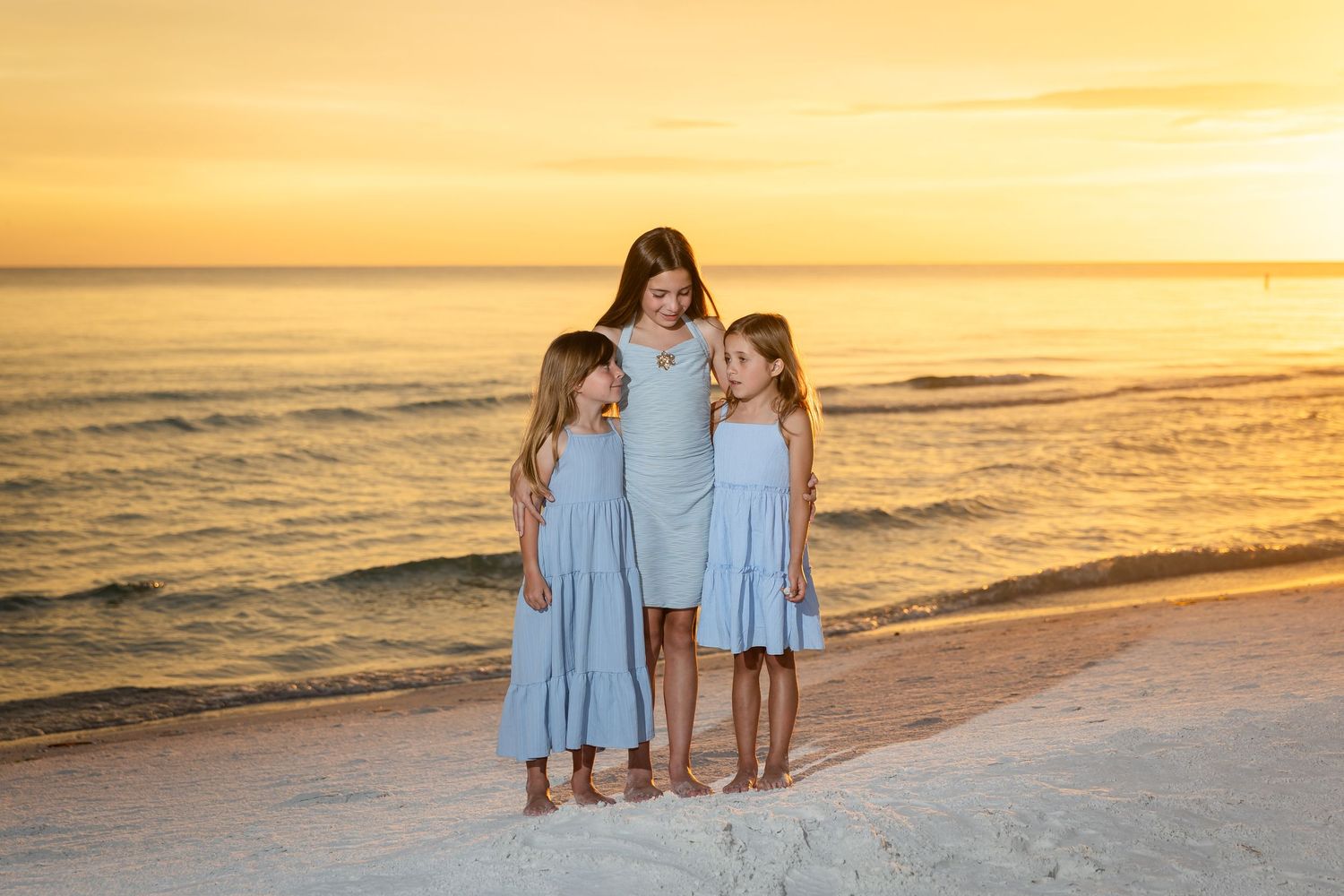 A mother and her two daughters in matching blue dresses pose on a white sand beach during a golden sunset.