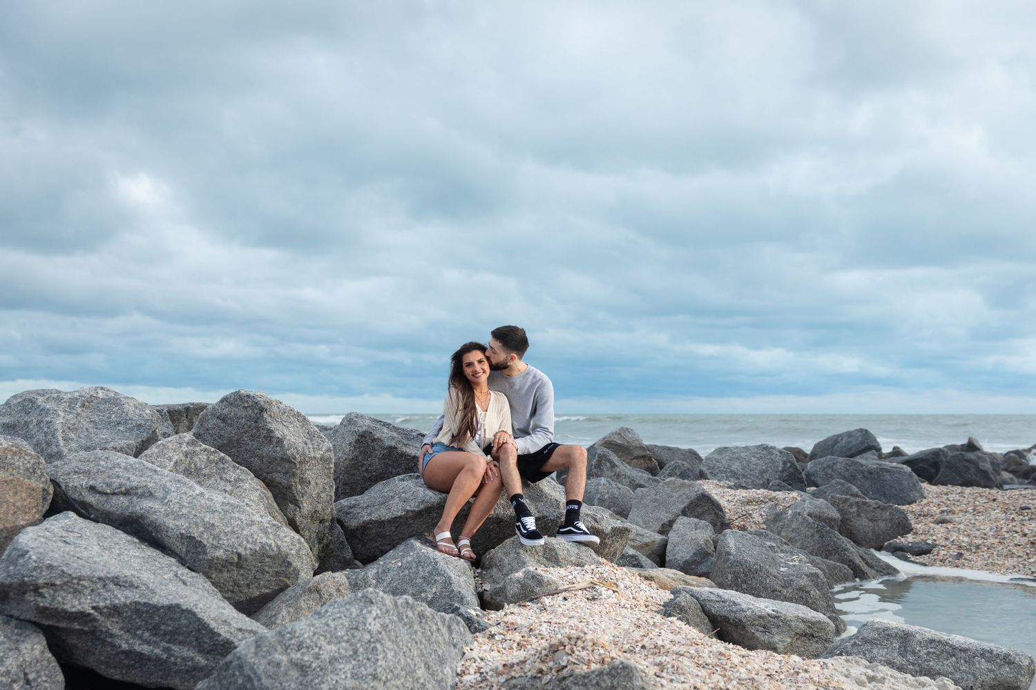 A couple sits romantically on rocky shoreline boulders under a cloudy sky with ocean waves in the background.