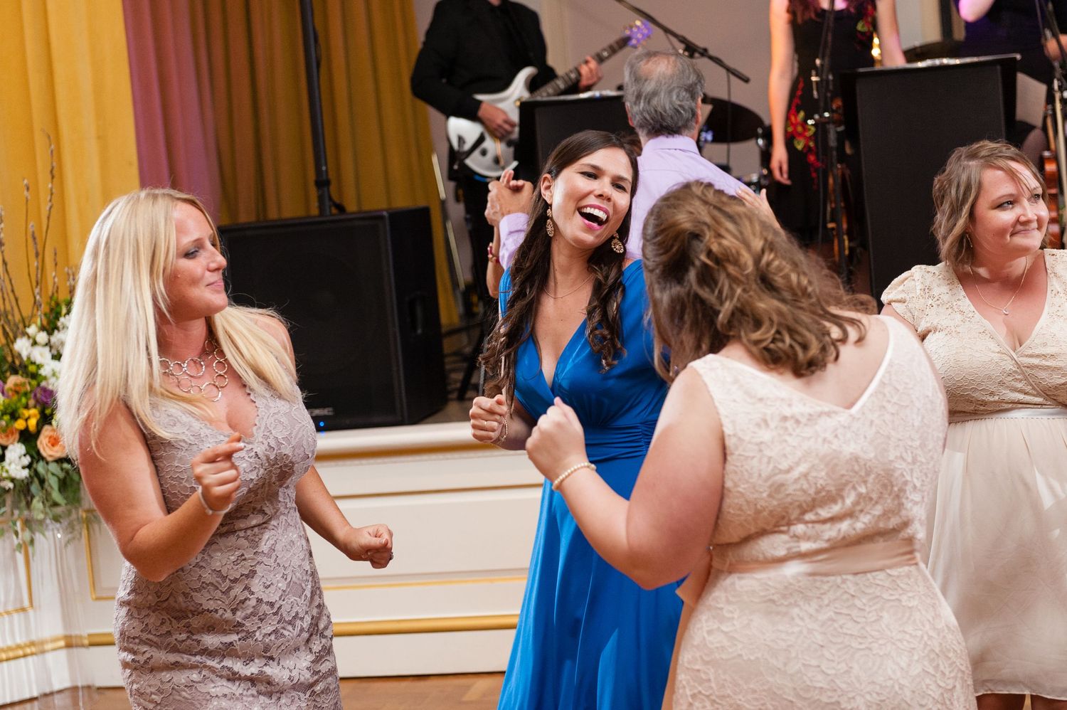 Women laugh and dance joyfully together at wedding reception with live band performing on stage.