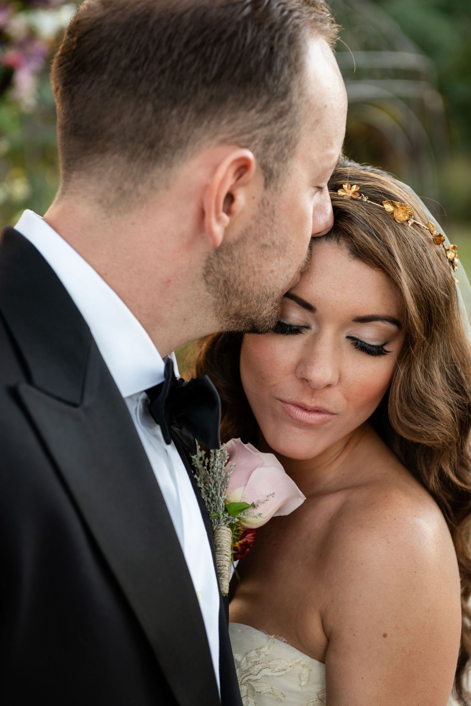 Groom in black tuxedo kissing bride on forehead, bride wearing gold floral crown and strapless lace gown.