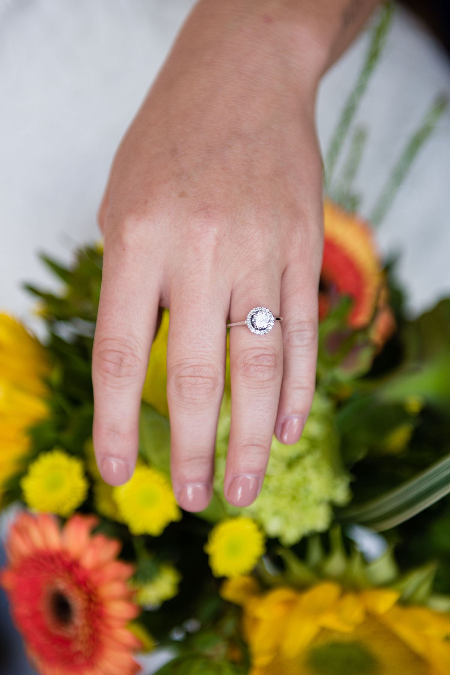 Close-up of a diamond halo engagement ring on a bride's hand over a colorful bouquet of yellow and orange flowers.