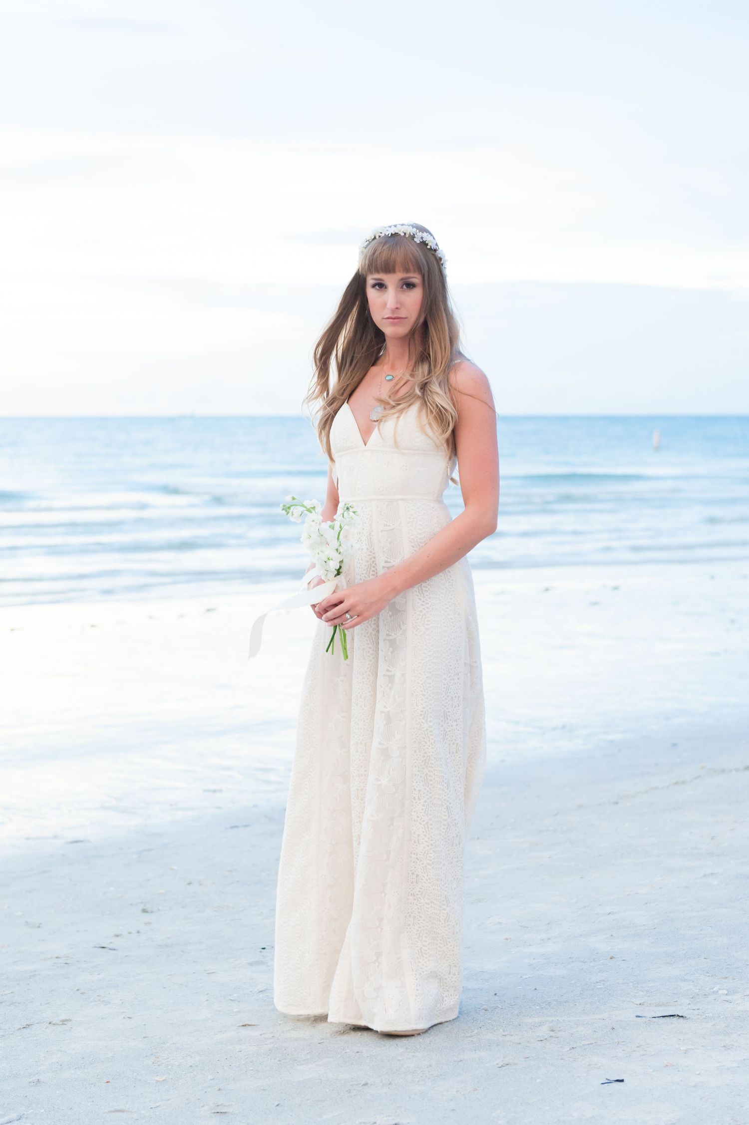Elegant bride in lace wedding gown holding white bouquet standing on sandy beach with ocean backdrop.