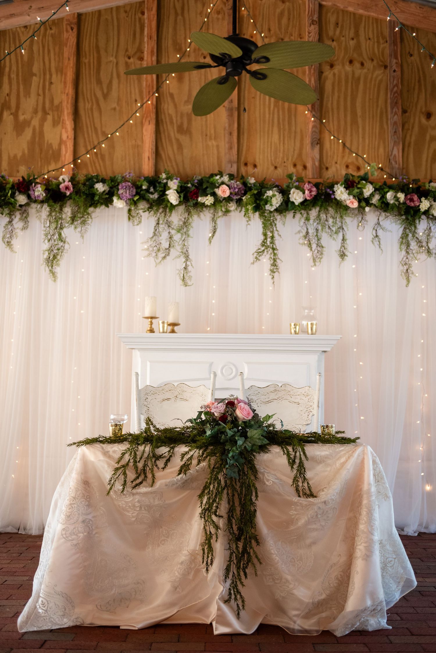 Elegant wedding sweetheart table with lush greenery, pink flowers, fairy lights, and white draping in a barn venue.