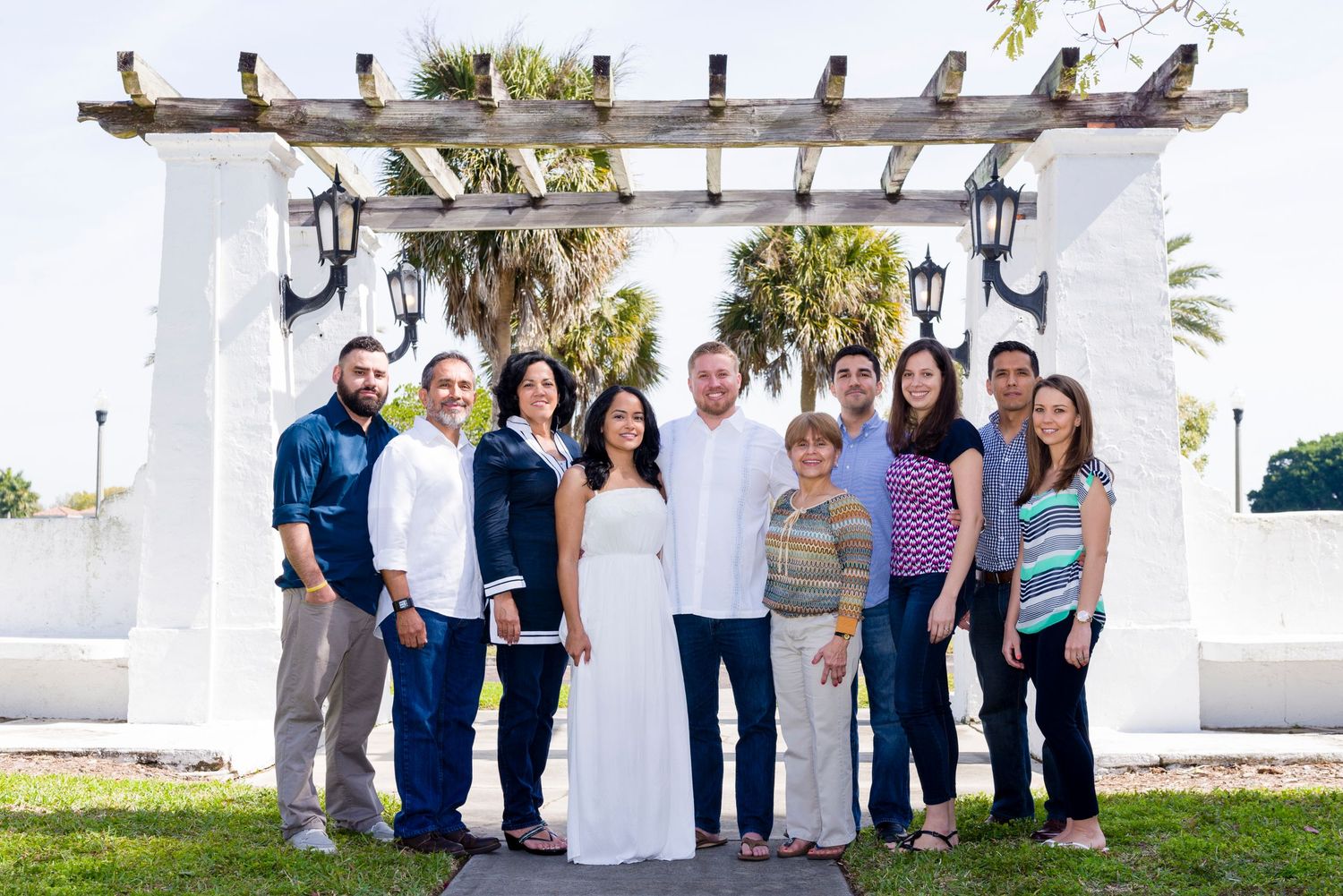 Group of eleven people smiling outdoors under a wooden pergola with palm trees, dressed in smart casual attire for a celebration.