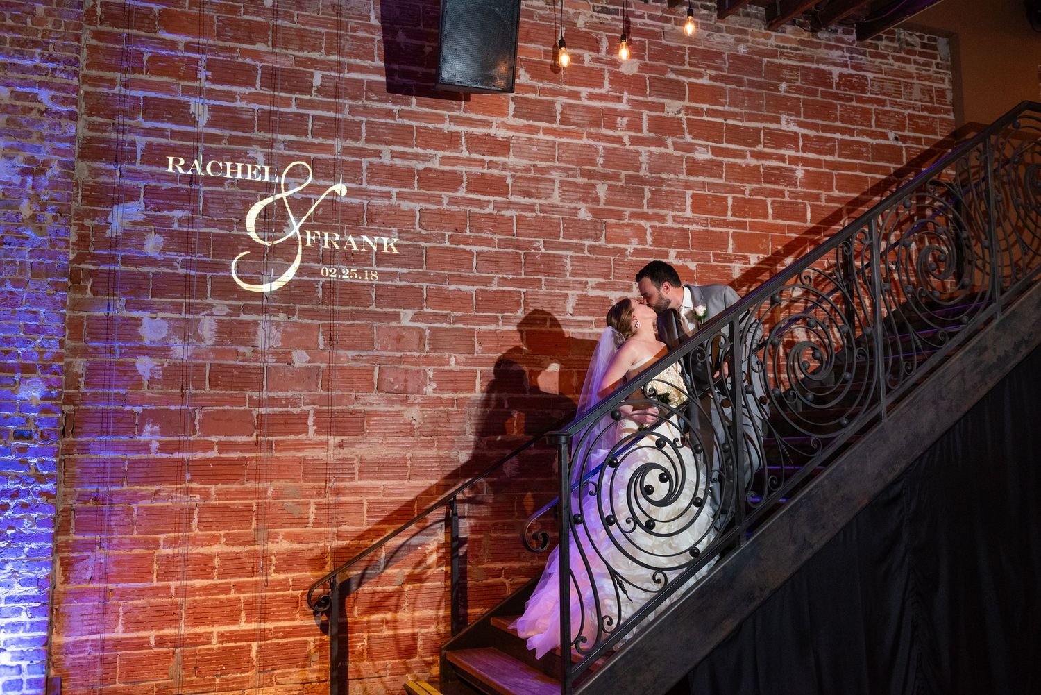 Bride and groom share a romantic kiss on ornate iron staircase with brick wall backdrop at Rachel and Ethan wedding.