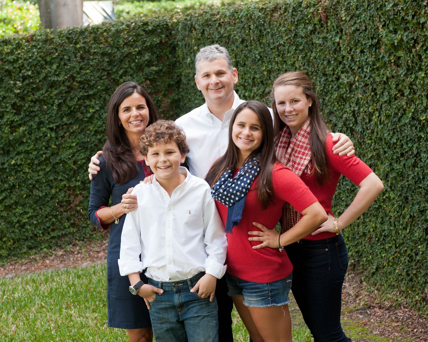 Happy family of five posing outdoors in front of lush green hedge, smiling together in casual attire.