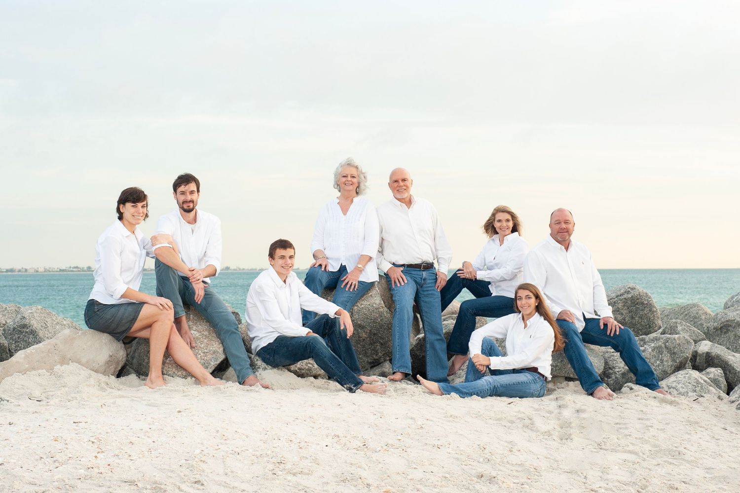 Family group photo on sandy beach, all wearing white shirts and jeans, posing on rocks with ocean in background.