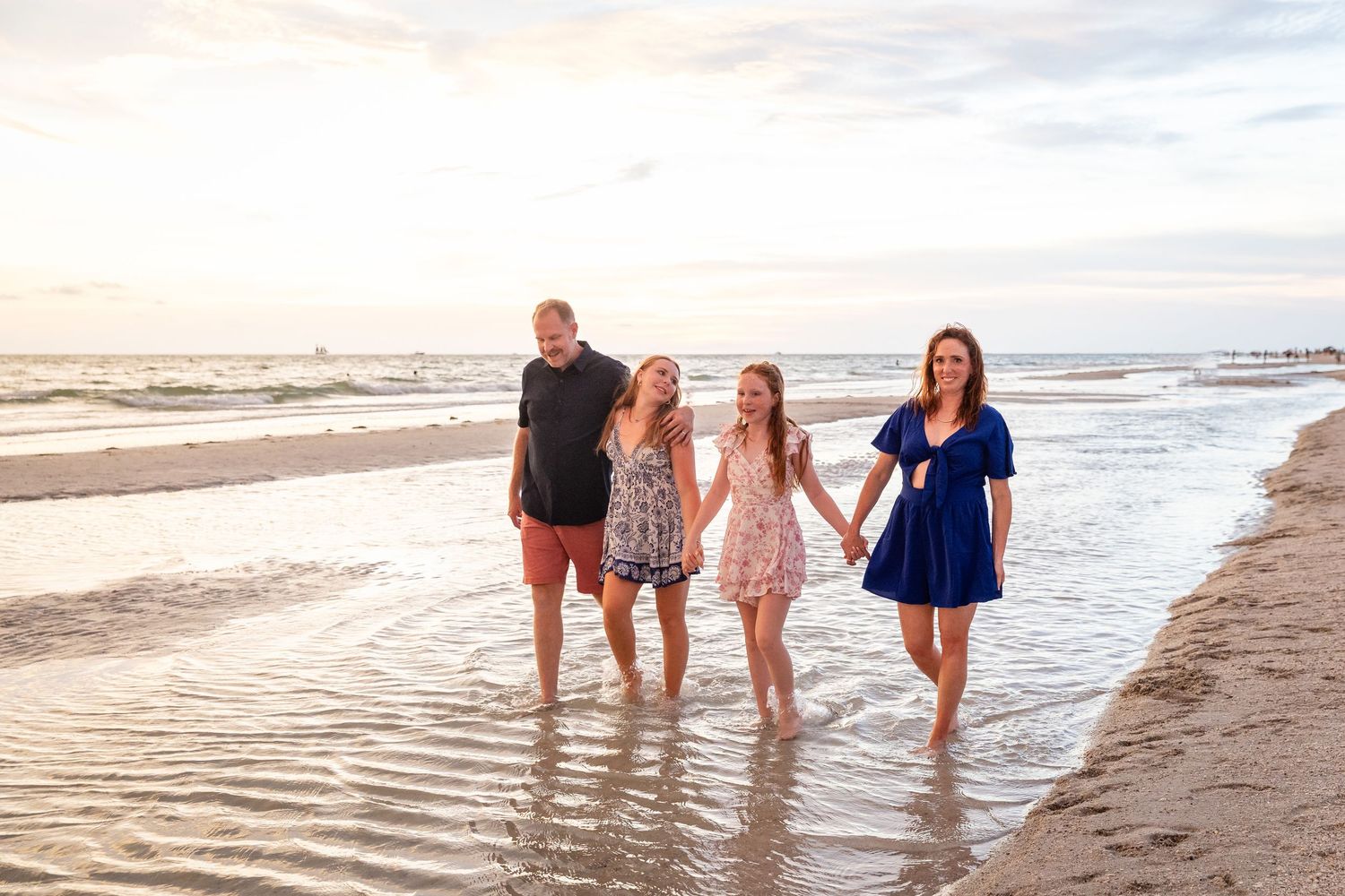 Family of four walking hand-in-hand on a sandy beach at sunset, smiling and enjoying a seaside vacation together.