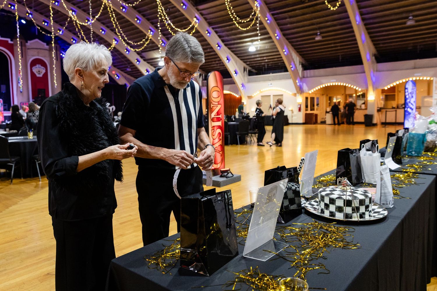 Two guests browse silent auction items on a decorated table at a casino-themed charity gala event.