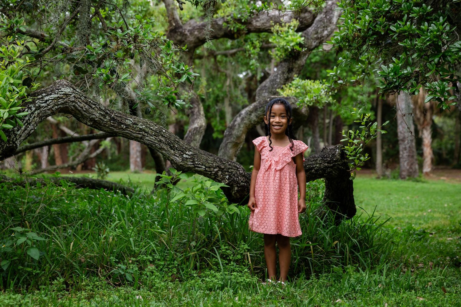 Young girl in pink dress smiling in lush green park with large twisted tree branches behind her.