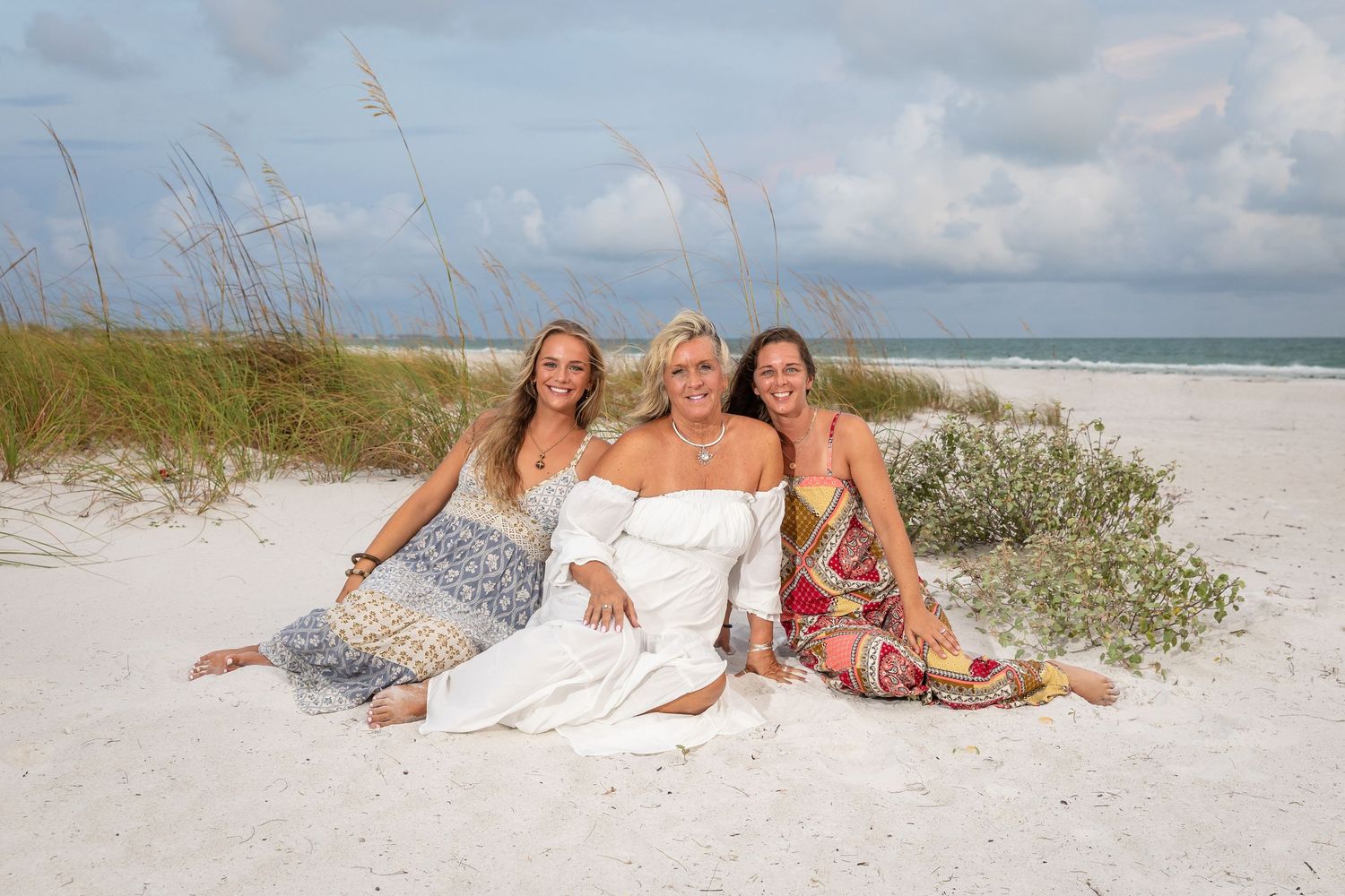Three women smile together on a white sandy beach with sea oats and ocean waves in the background.