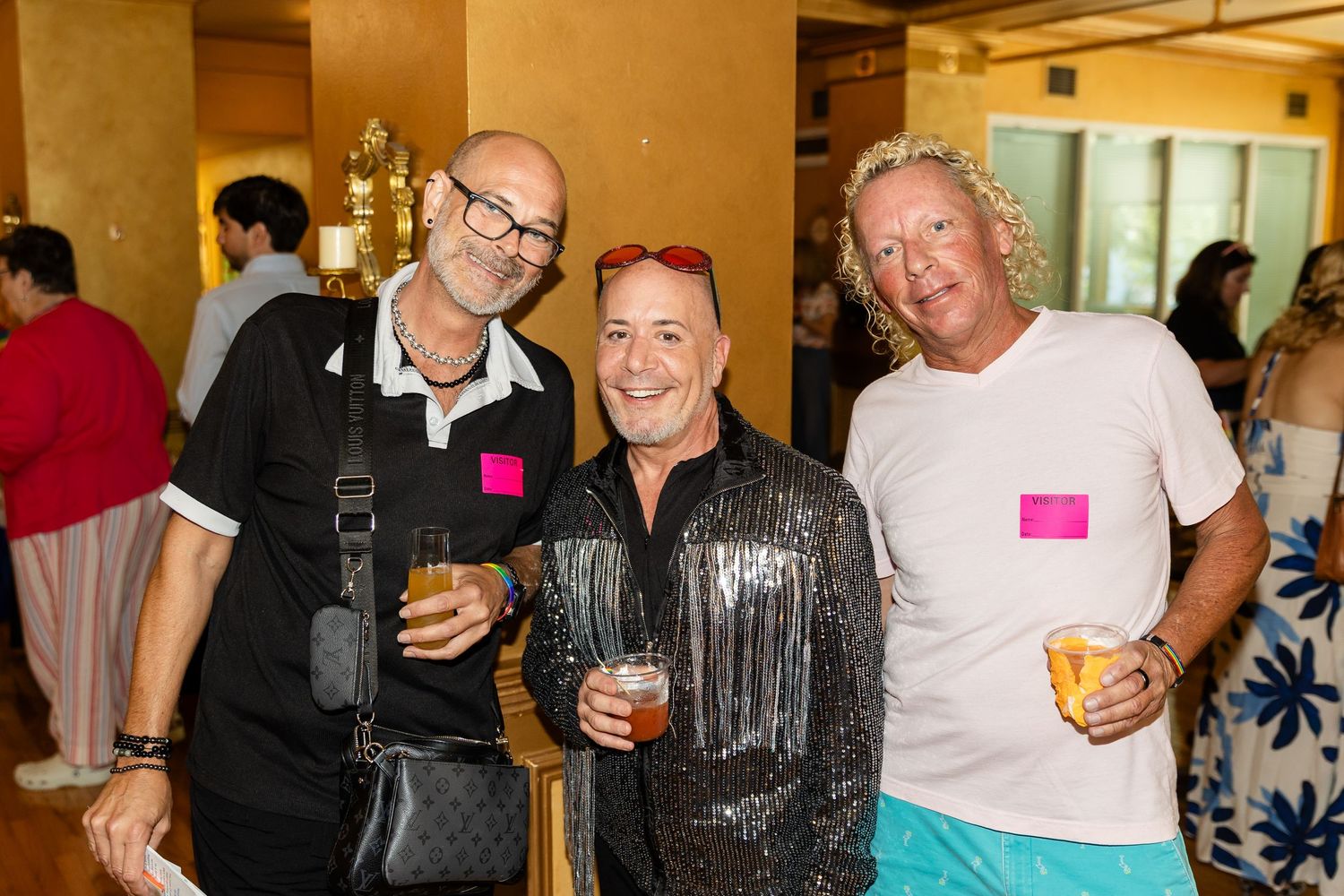 Three smiling men posing together at a social event, one wearing a silver sequin jacket, holding drinks.