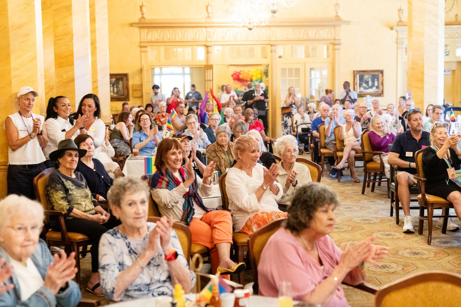 Large group of seniors applauding and celebrating together at a colorful indoor Pride community gathering event.