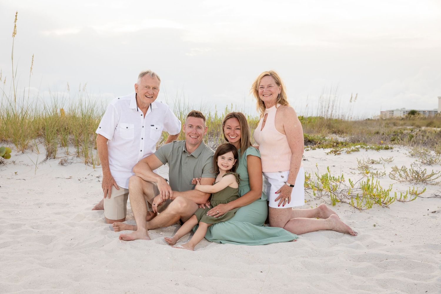 Family of five poses on sandy beach with sea oats, smiling warmly at sunset in casual summer attire.