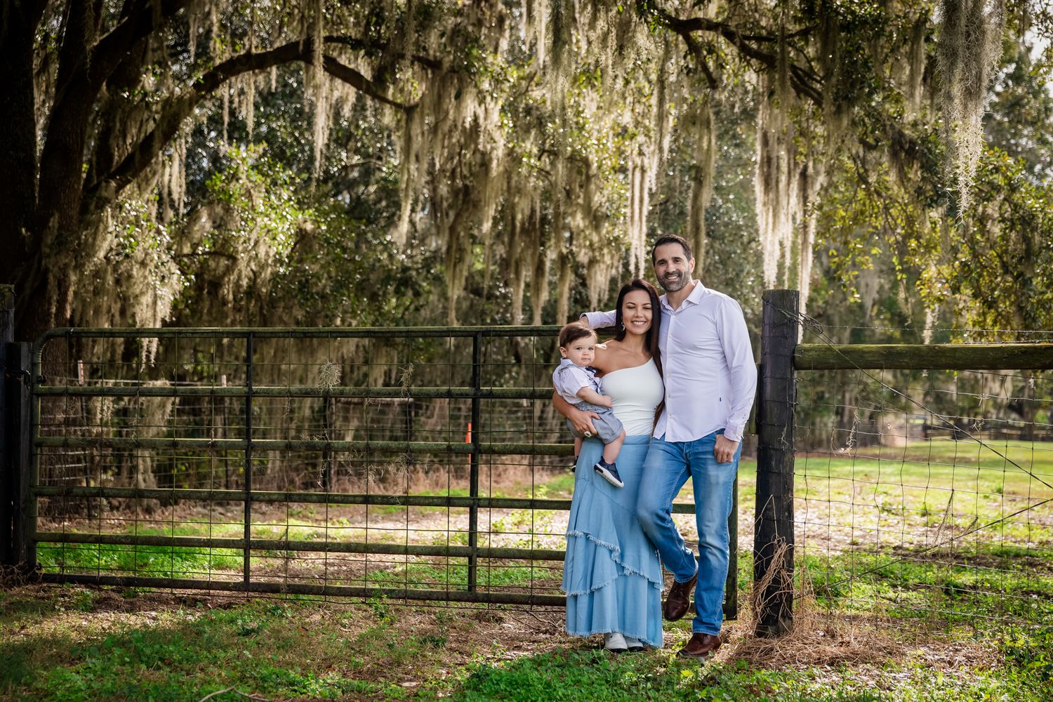 A happy family of three poses by a rustic black gate under sprawling Spanish moss-draped oak trees.