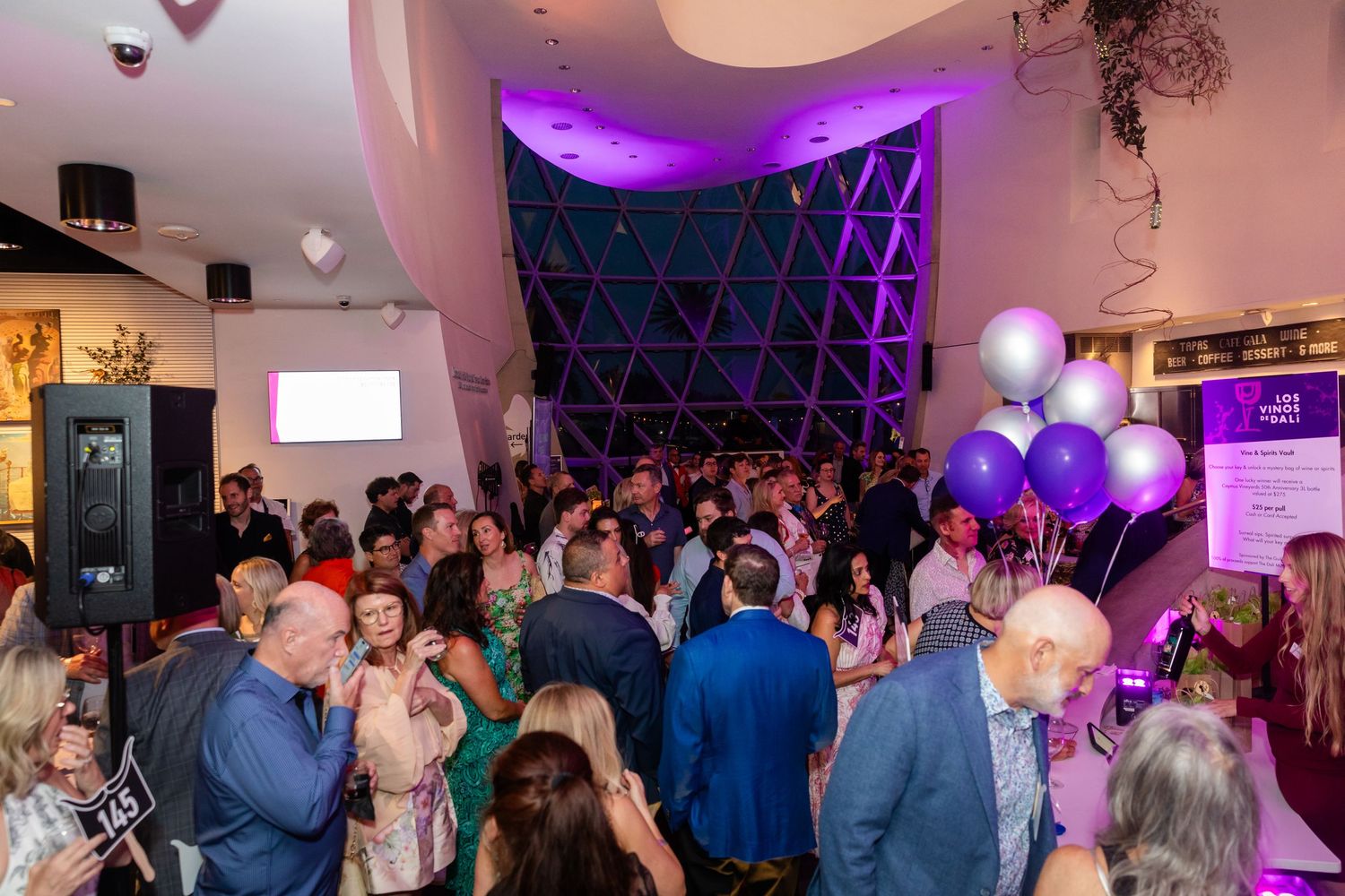 Crowded indoor party venue with purple balloons, ambient lighting, and guests mingling together.