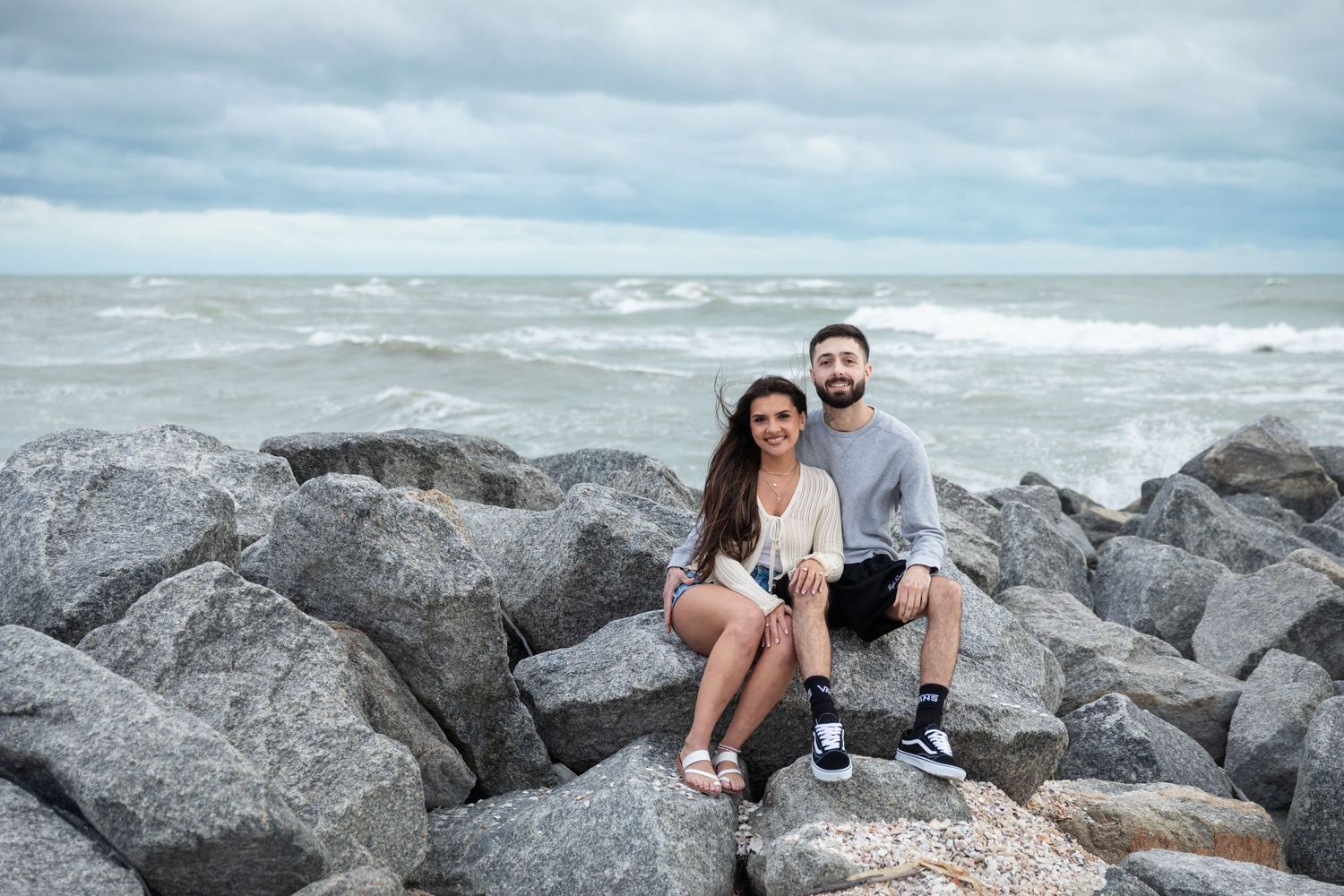 A couple sits smiling on rocky shoreline with choppy ocean waves and cloudy sky behind them.