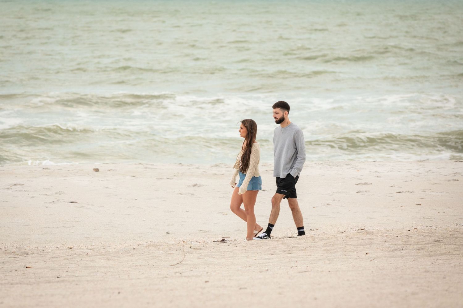 Couple walking together on a sandy beach with ocean waves in the background on a cloudy day.