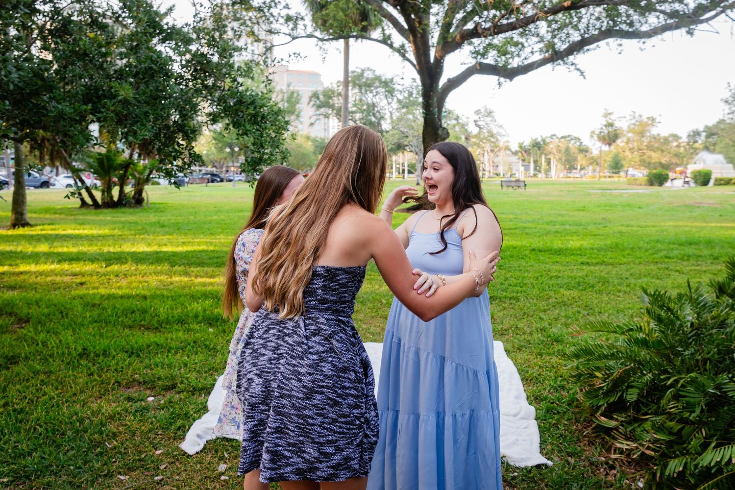 Three young women laughing and embracing outdoors on green grass near trees during a sunny day.
