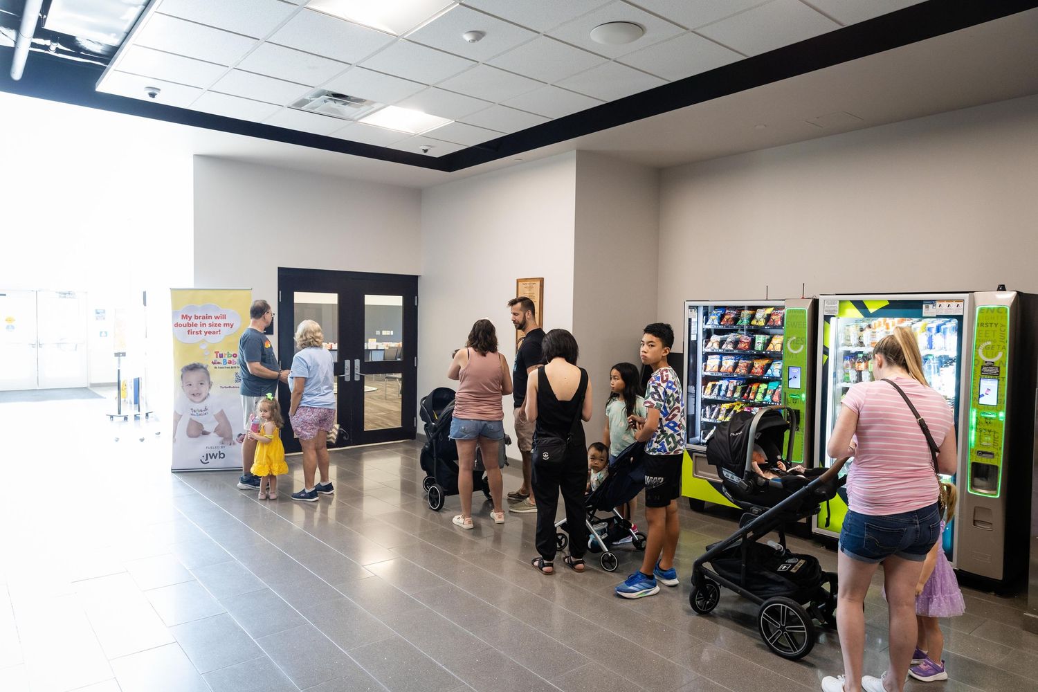 People queue near vending machines in a modern building lobby, with a stroller and promotional banner visible.