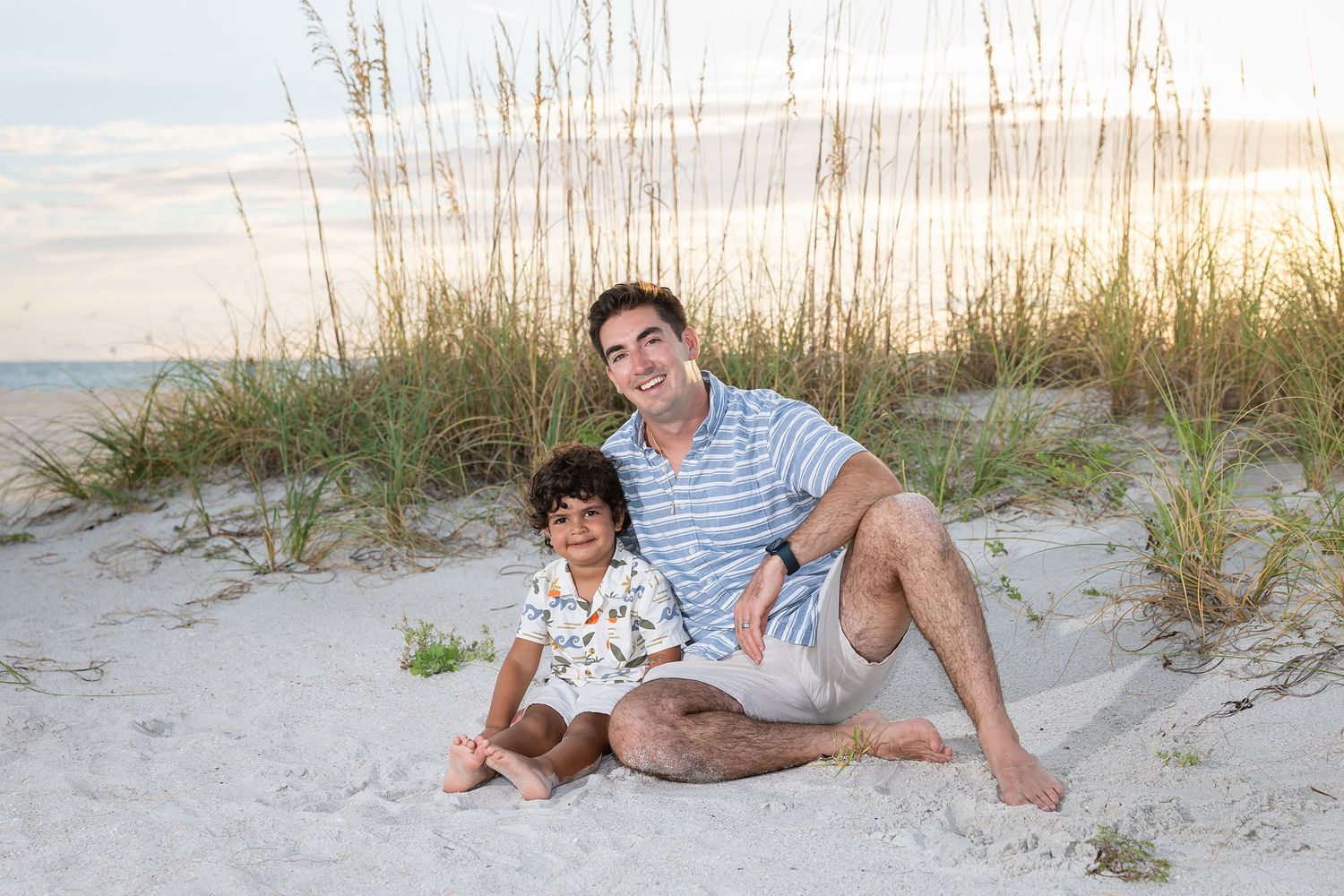 A man and young boy smile together on a sandy beach at sunset, sitting among sea oats and dunes.