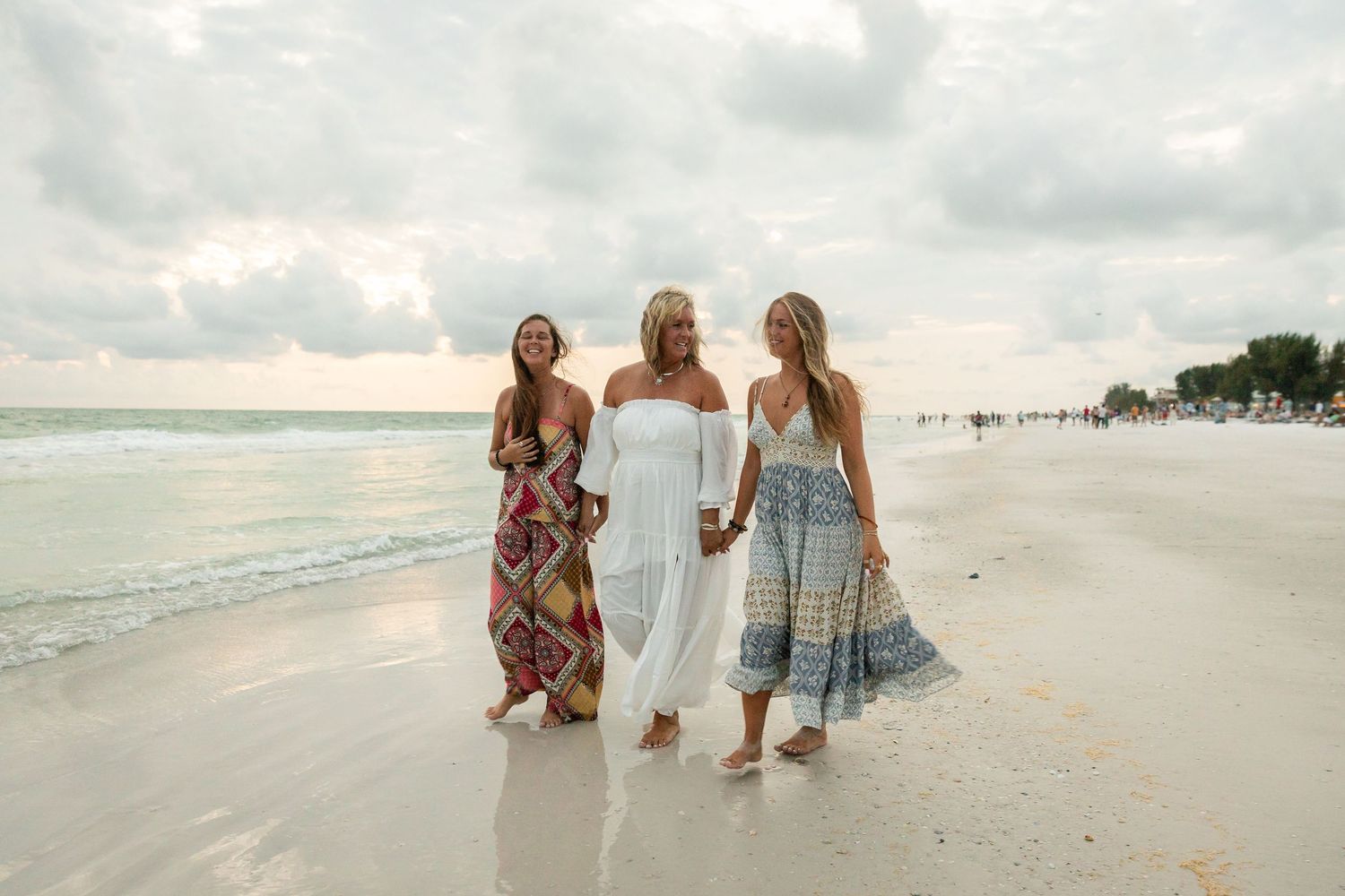 Three women in colorful summer dresses walking along a sandy beach with ocean waves and cloudy sky.