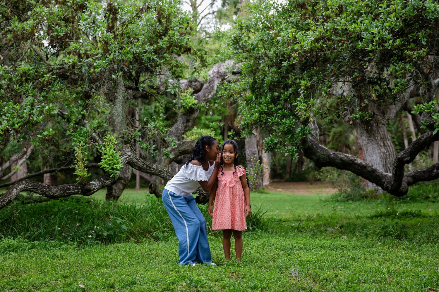 An older girl leans in to whisper to a younger girl in a pink dress among lush green trees.