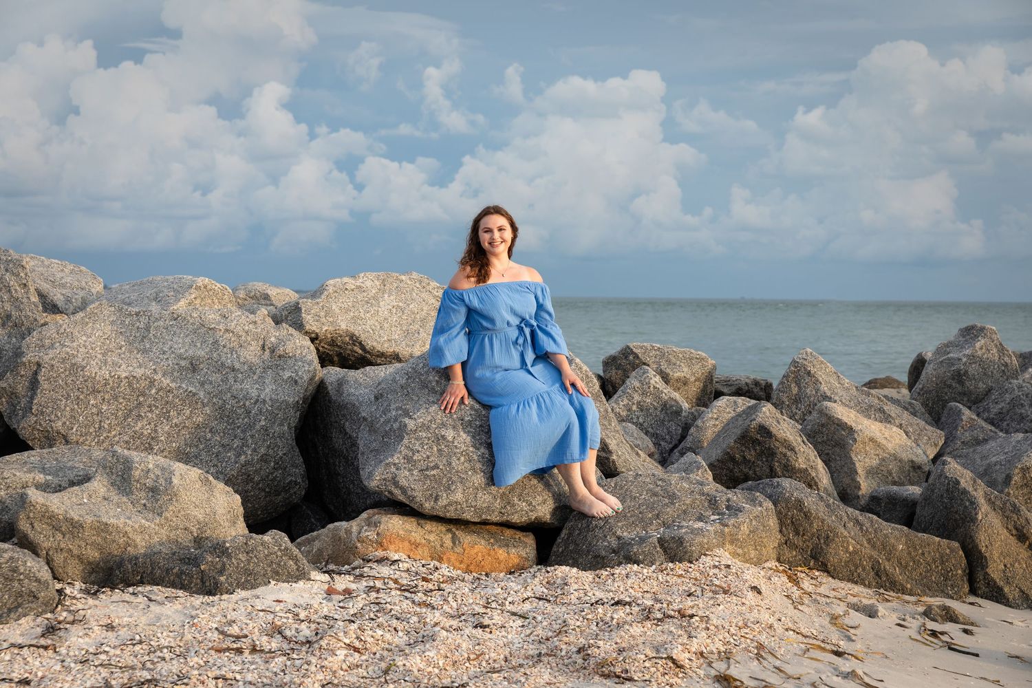 Woman in blue off-shoulder dress sitting on rocky beach with ocean and cloudy sky backdrop.