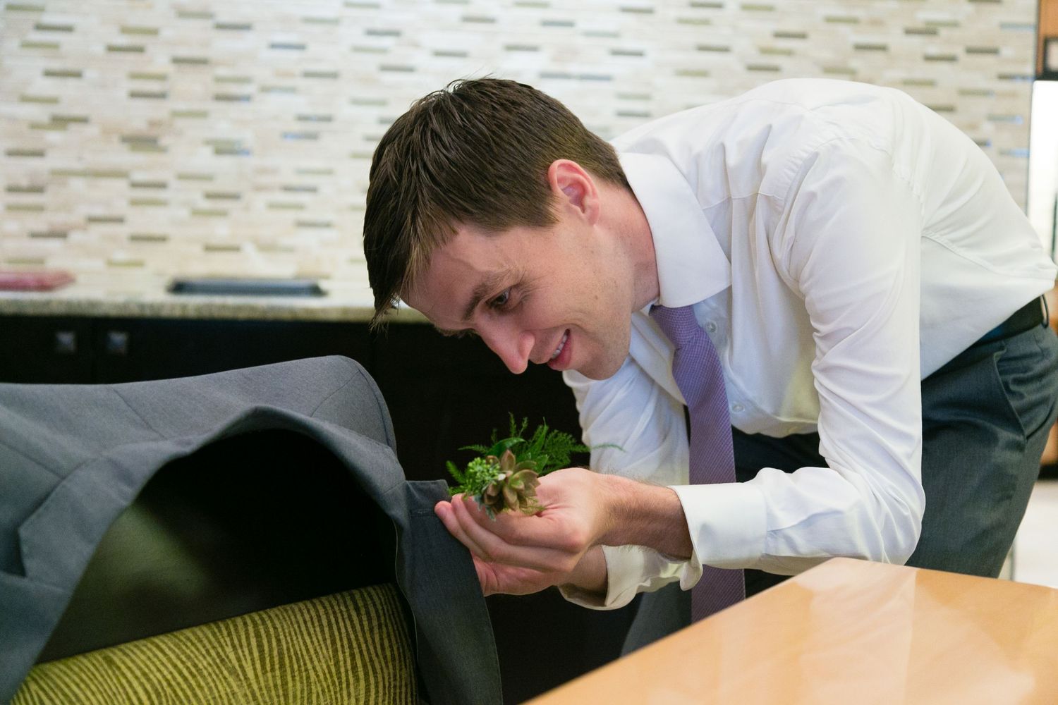 A smiling man in a white shirt and tie leaning over to smell a small bouquet of greenery.