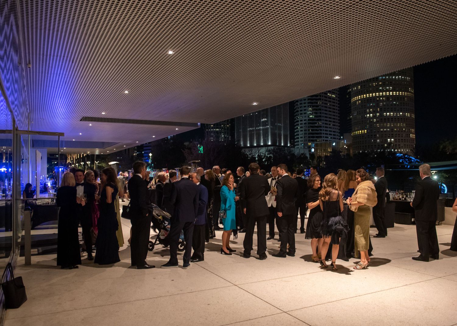 Elegantly dressed guests mingle on an outdoor terrace at a formal evening gala with city skyline views.