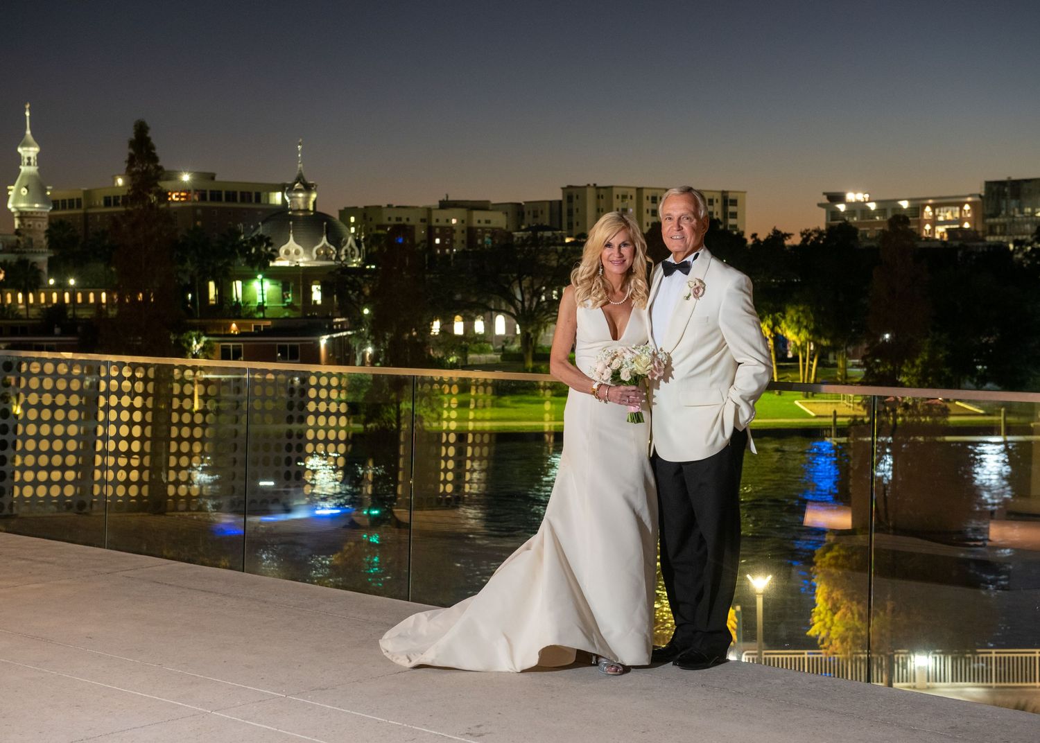 Bride and groom pose on rooftop terrace at night with city skyline lights reflecting on water below.
