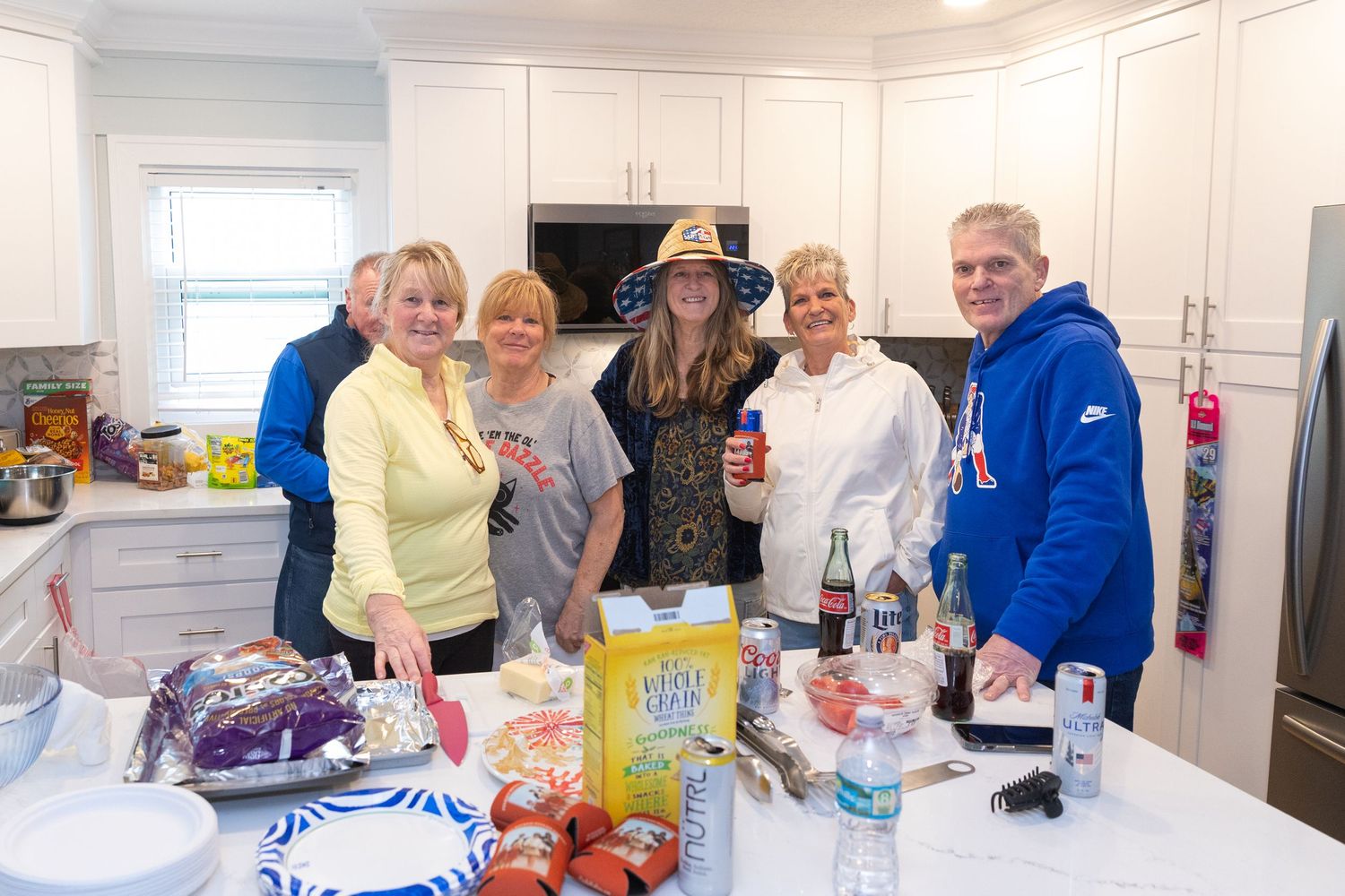 Group of six people gathered around a kitchen island filled with food and drinks at a party.