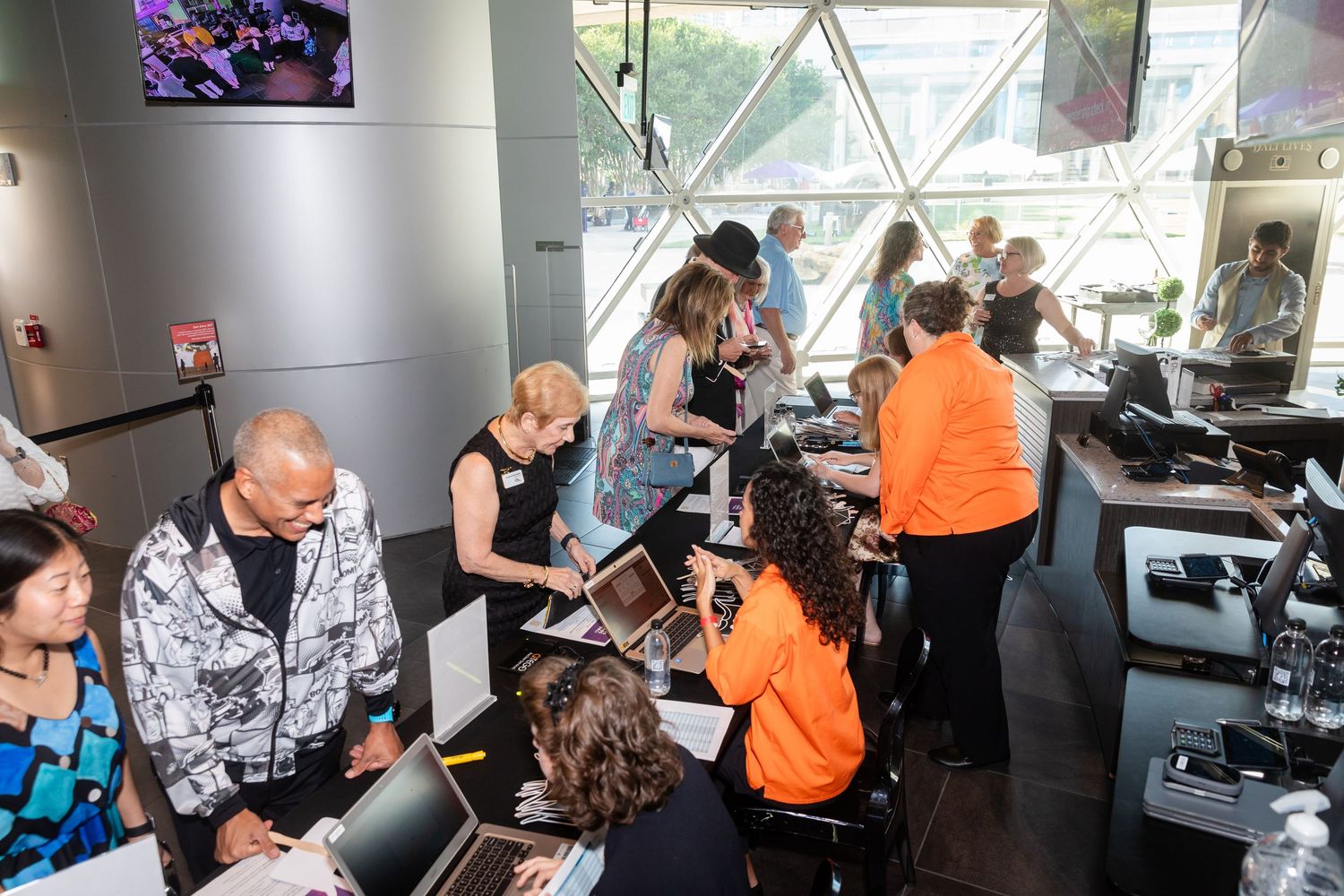Busy event registration desk with attendees checking in, staff in orange shirts assisting guests inside a modern venue.