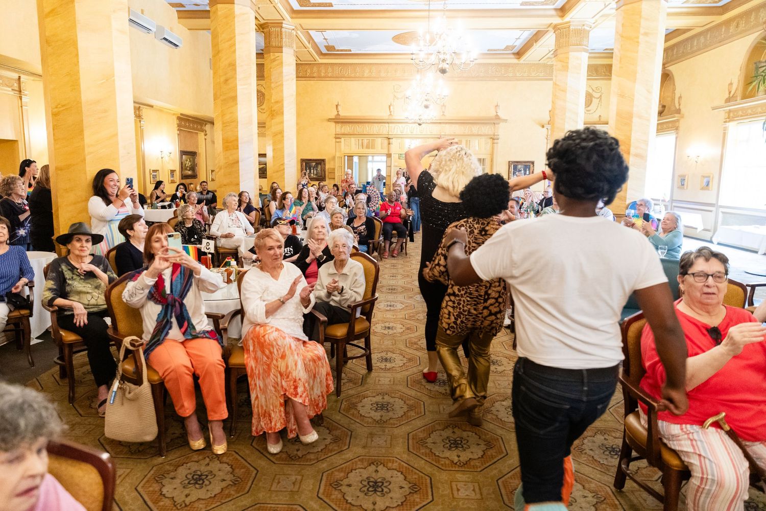 A lively fashion show audience applauds as a model walks through a grand ornate ballroom.