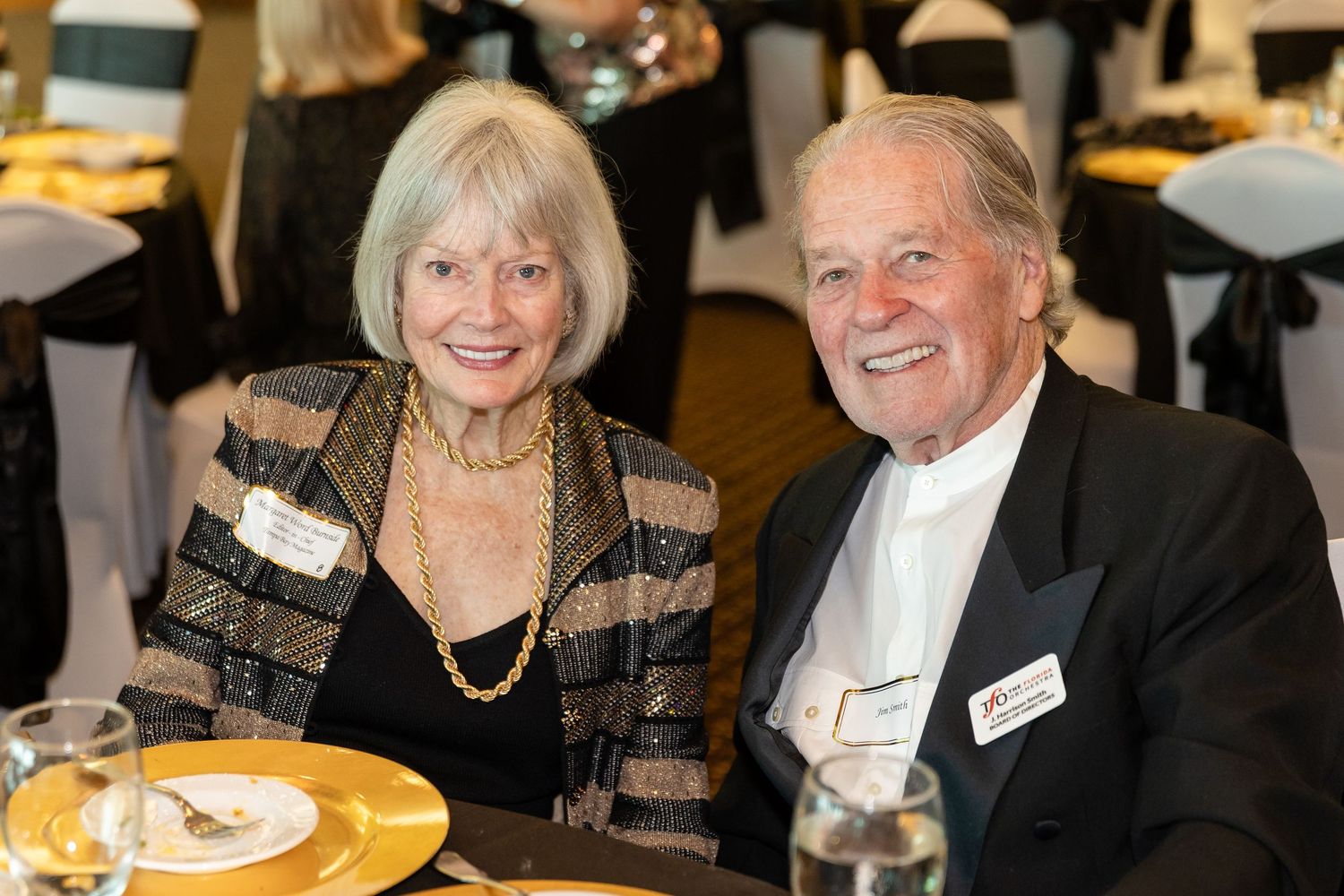 Smiling senior couple in formal attire seated at a gold-themed gala dinner event table.