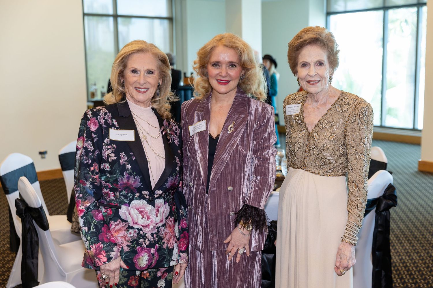 Three elegantly dressed women posing together at a formal event in a banquet hall setting.