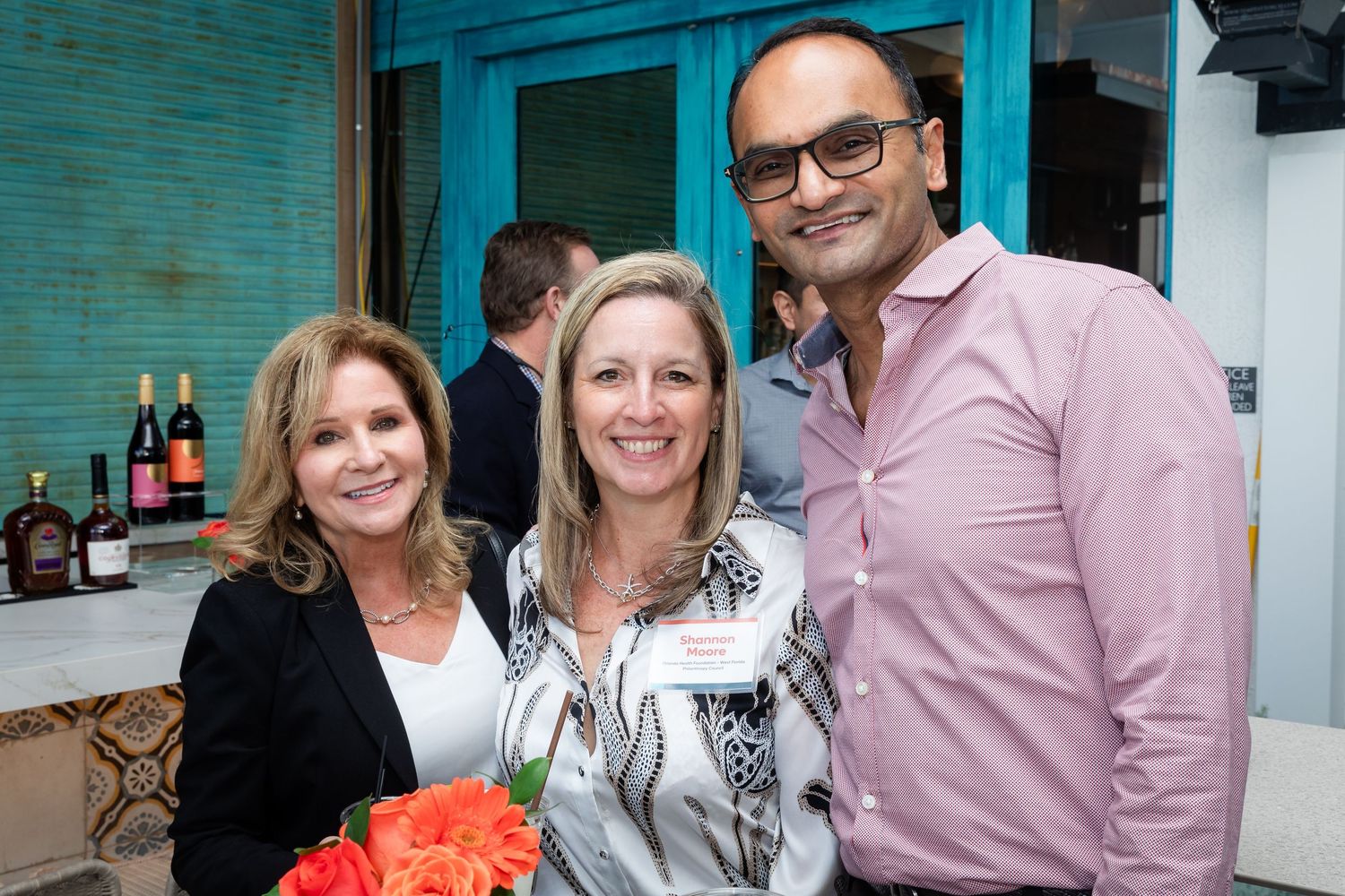 Three smiling professionals pose together at a casual networking event with liquor bottles visible in the background.