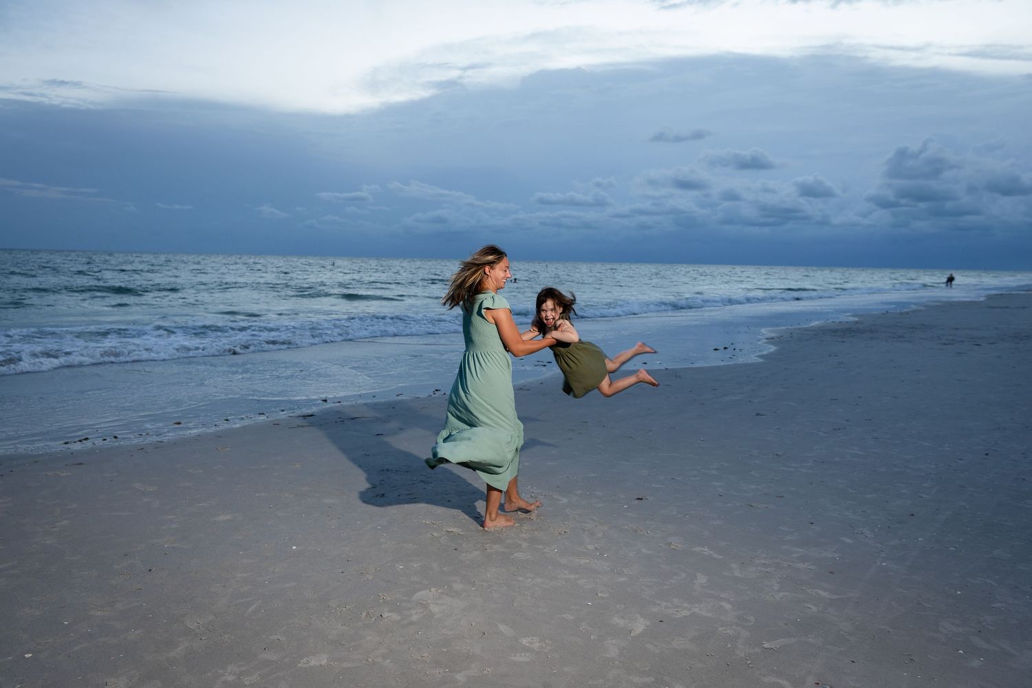 Woman in green dress swinging a child on a sandy beach at dusk with ocean waves in the background.