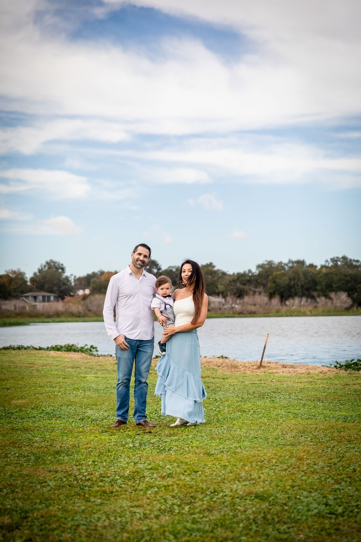 Happy family with baby posing by a lake on a sunny day, mother in blue dress, father in white shirt.