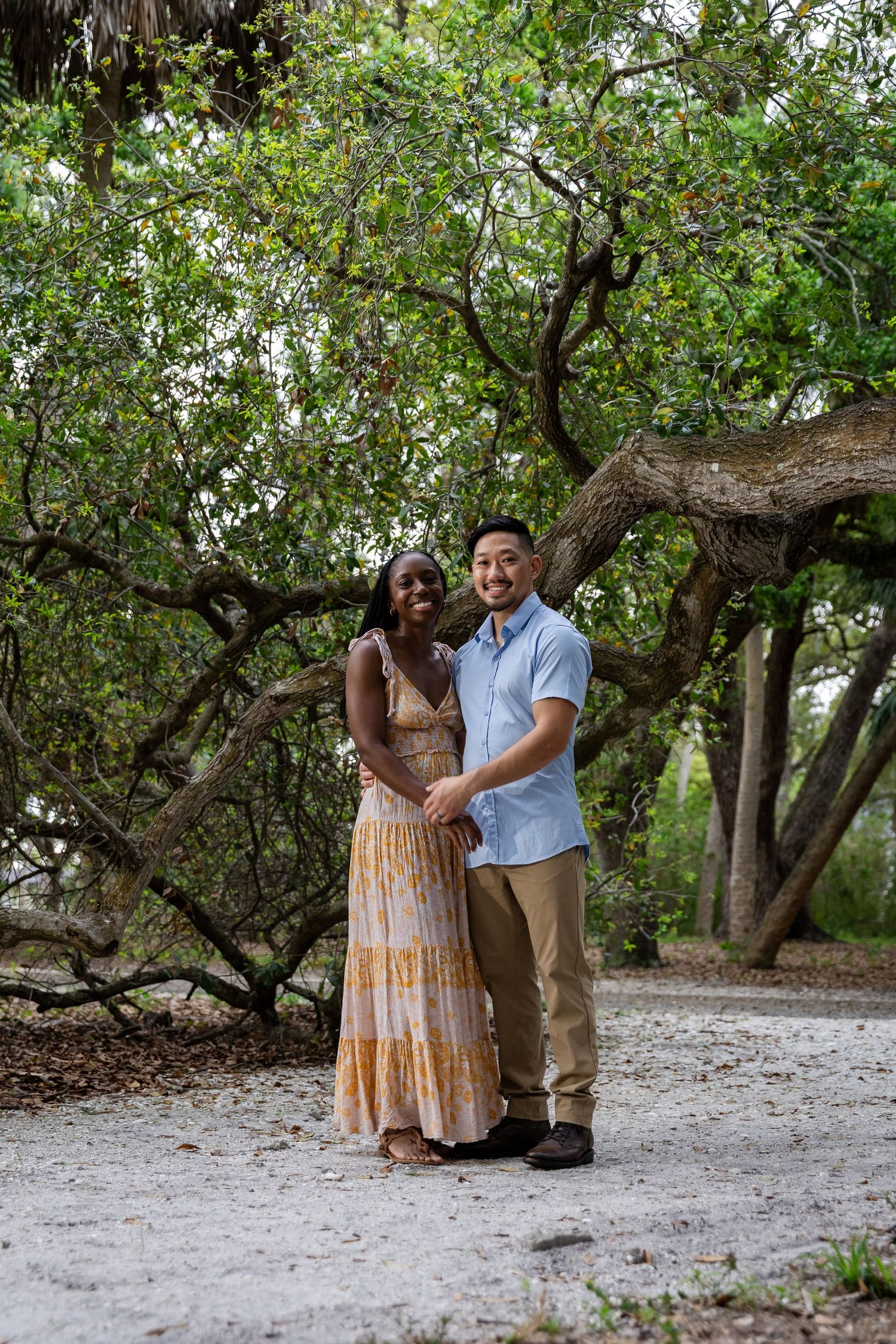 Smiling interracial couple embracing under a large tree on a sandy path in a natural outdoor setting.