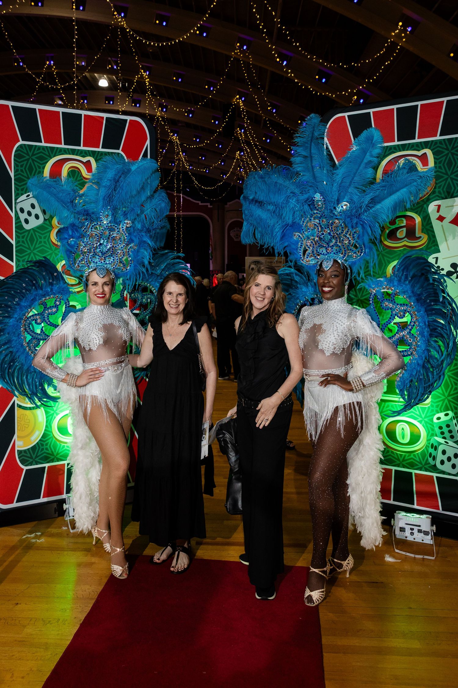 Two women in elaborate blue feathered showgirl costumes pose with guests on a red carpet at a casino-themed event.