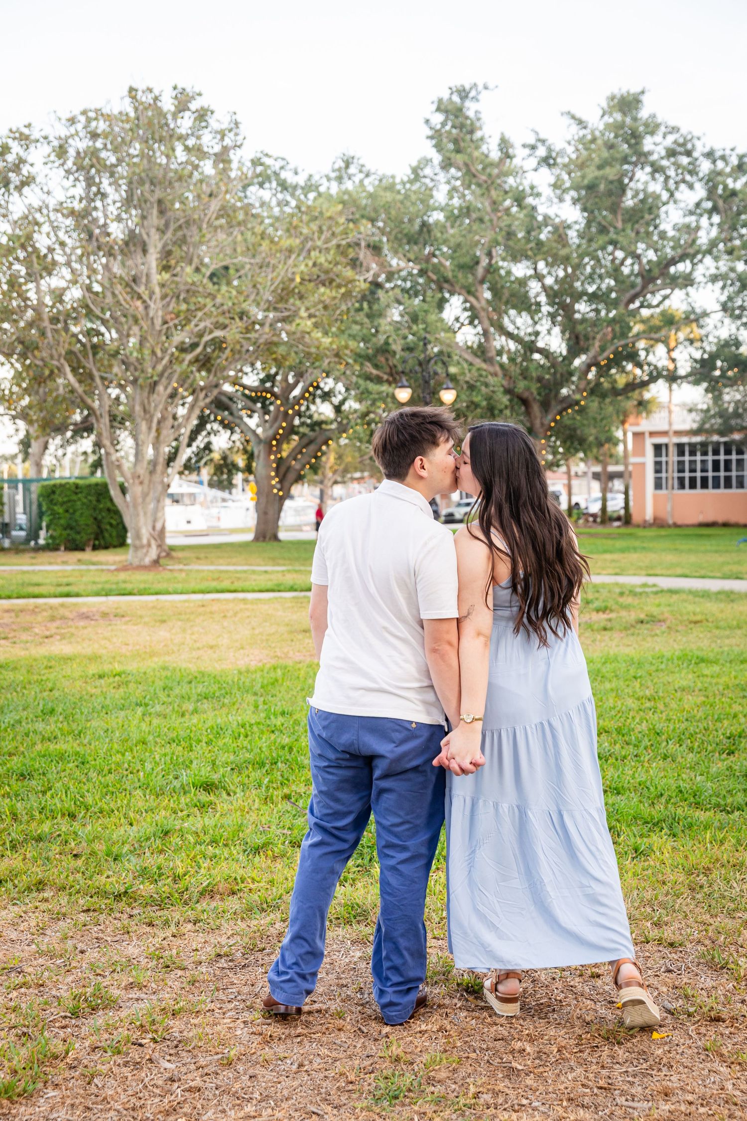 A couple holds hands and shares a kiss outdoors on green grass with large oak trees in the background.