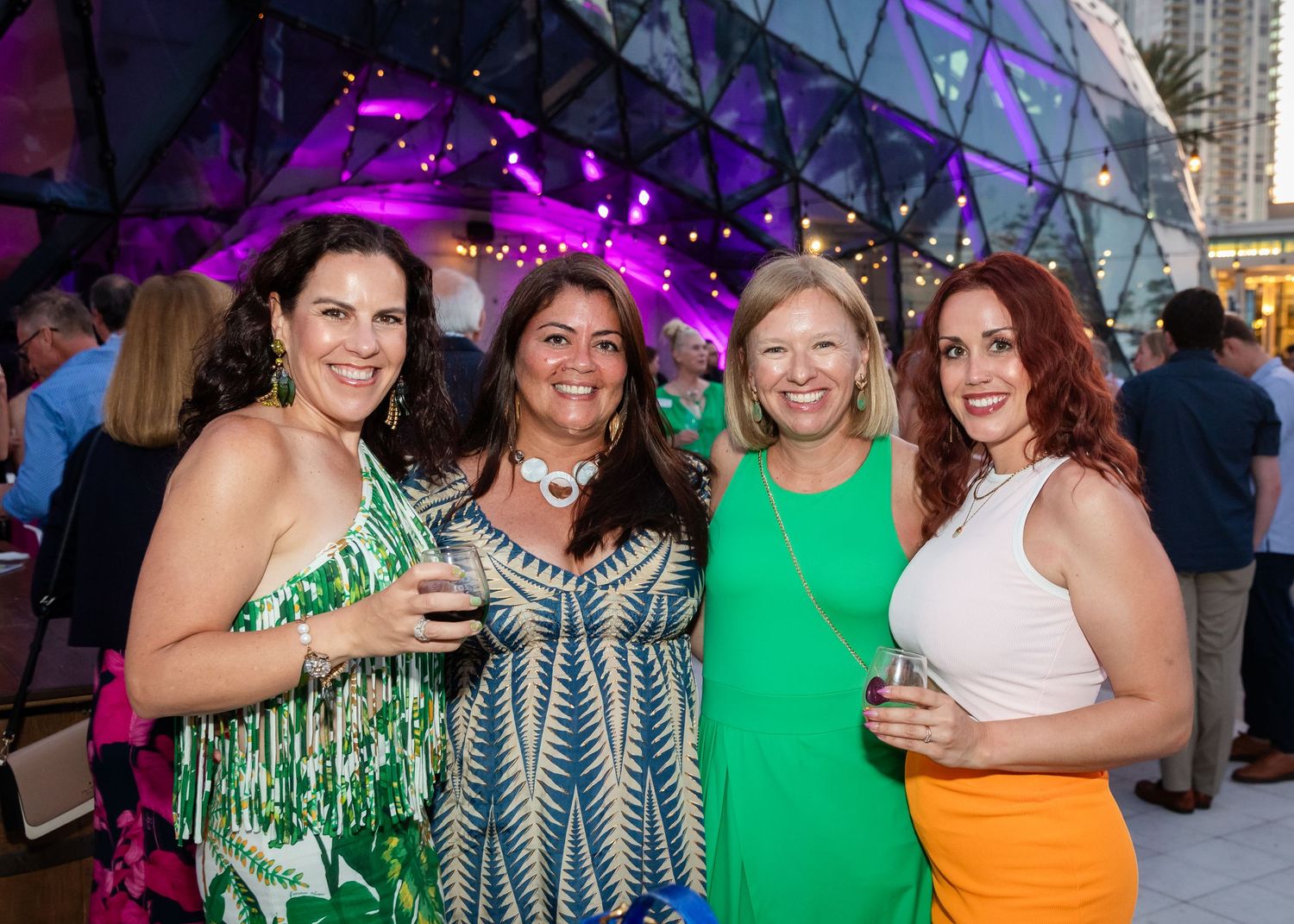 Four women smiling and holding drinks at an outdoor evening event near a modern dome-shaped venue.